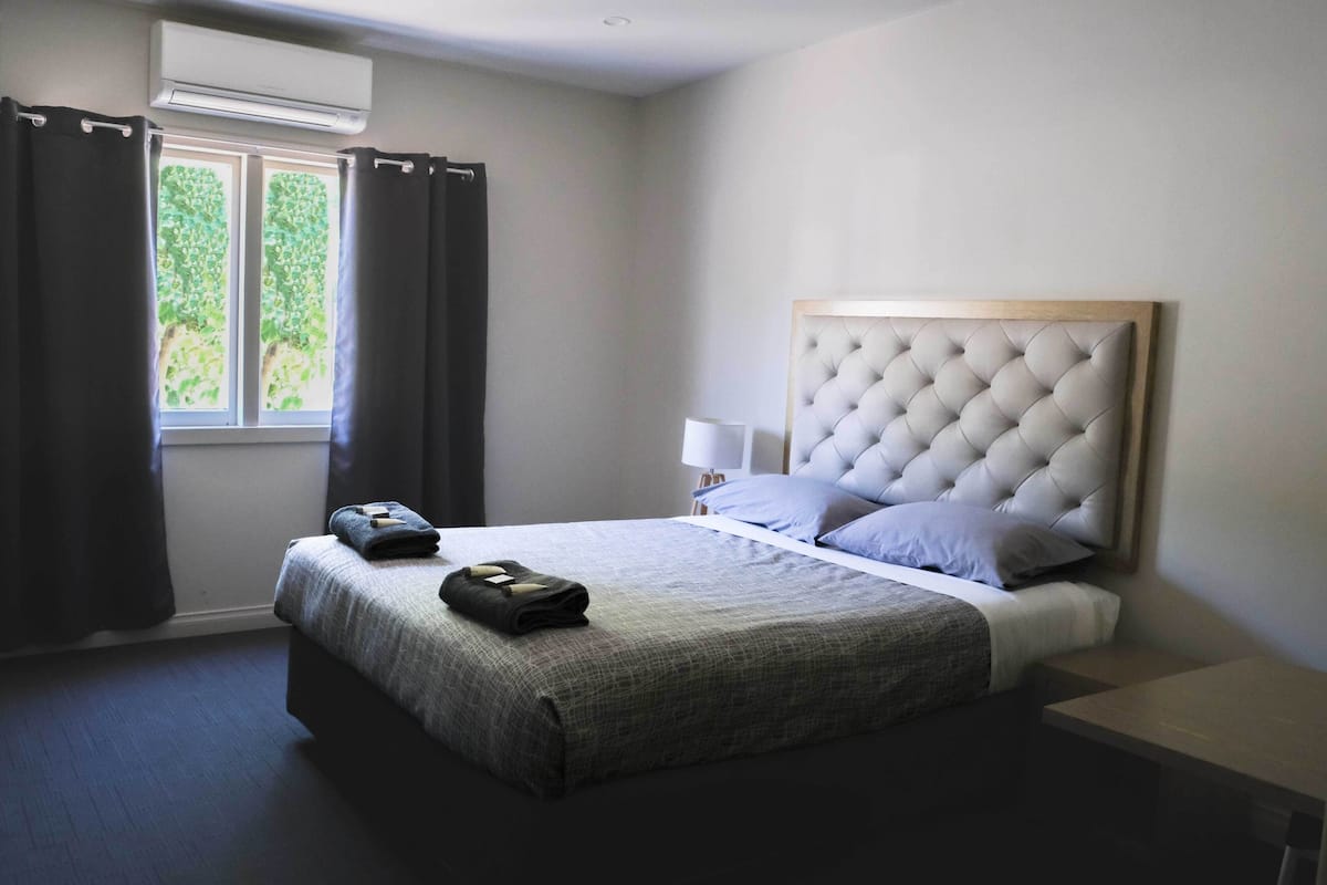 A bedroom with a large bed, a headboard, a bedside table, a lamp, and a window with curtains.