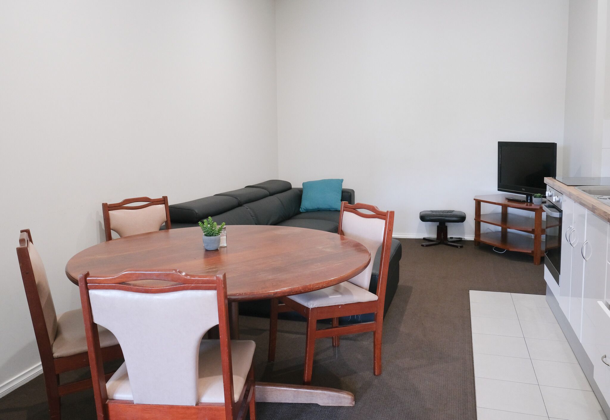 Un espace de vie compact comprenant une table de salle à manger ronde en bois, quatre chaises, un canapé, un petit téléviseur posé sur un meuble et un coin cuisine.