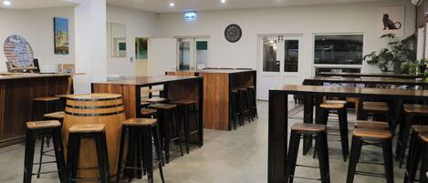Amplia zona de bar con taburetes de madera y una barra central, con un reloj en la pared y un panel de menú a la izquierda.