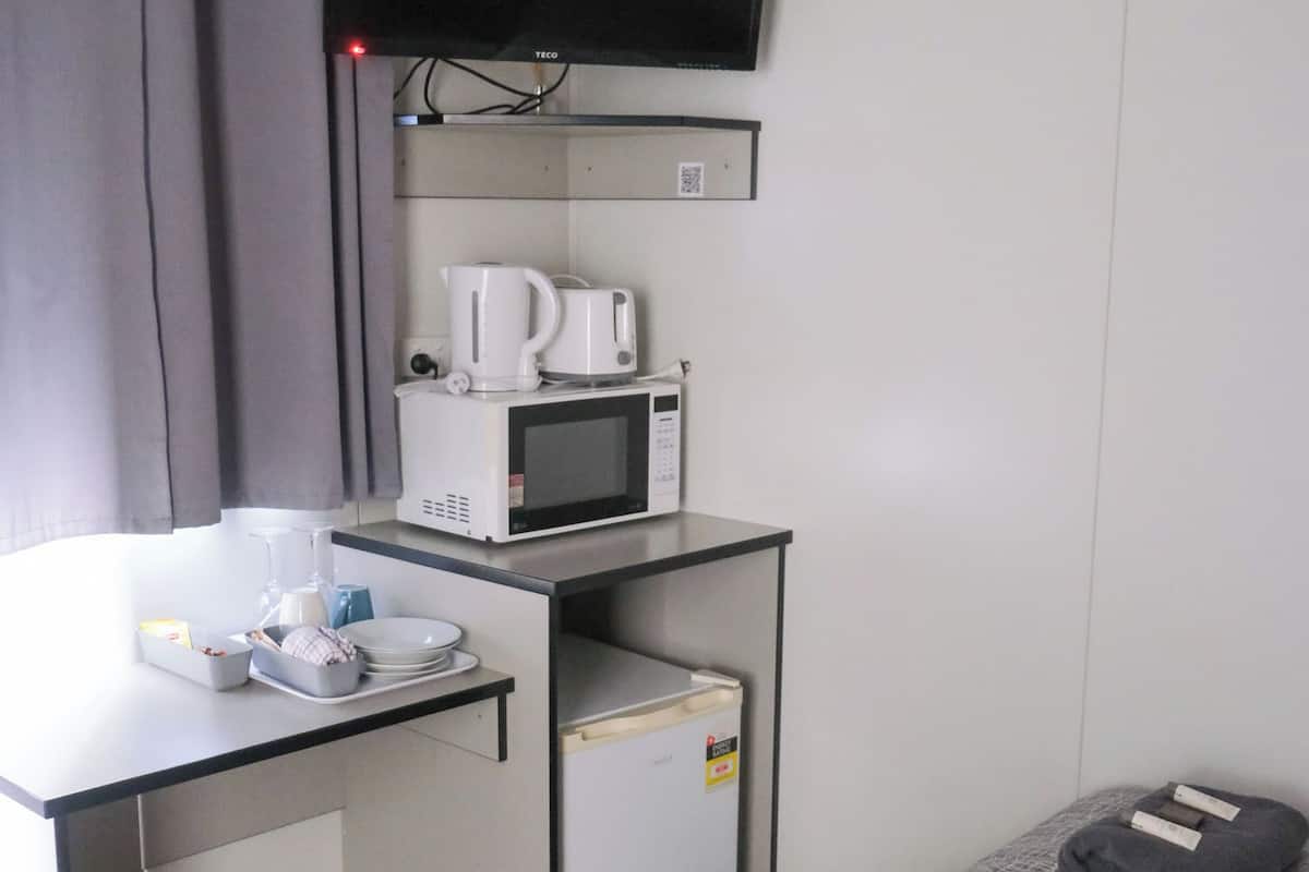 A compact hotel room with a kitchenette, a bed with a grey blanket, a TV mounted on the wall, and a small refrigerator.