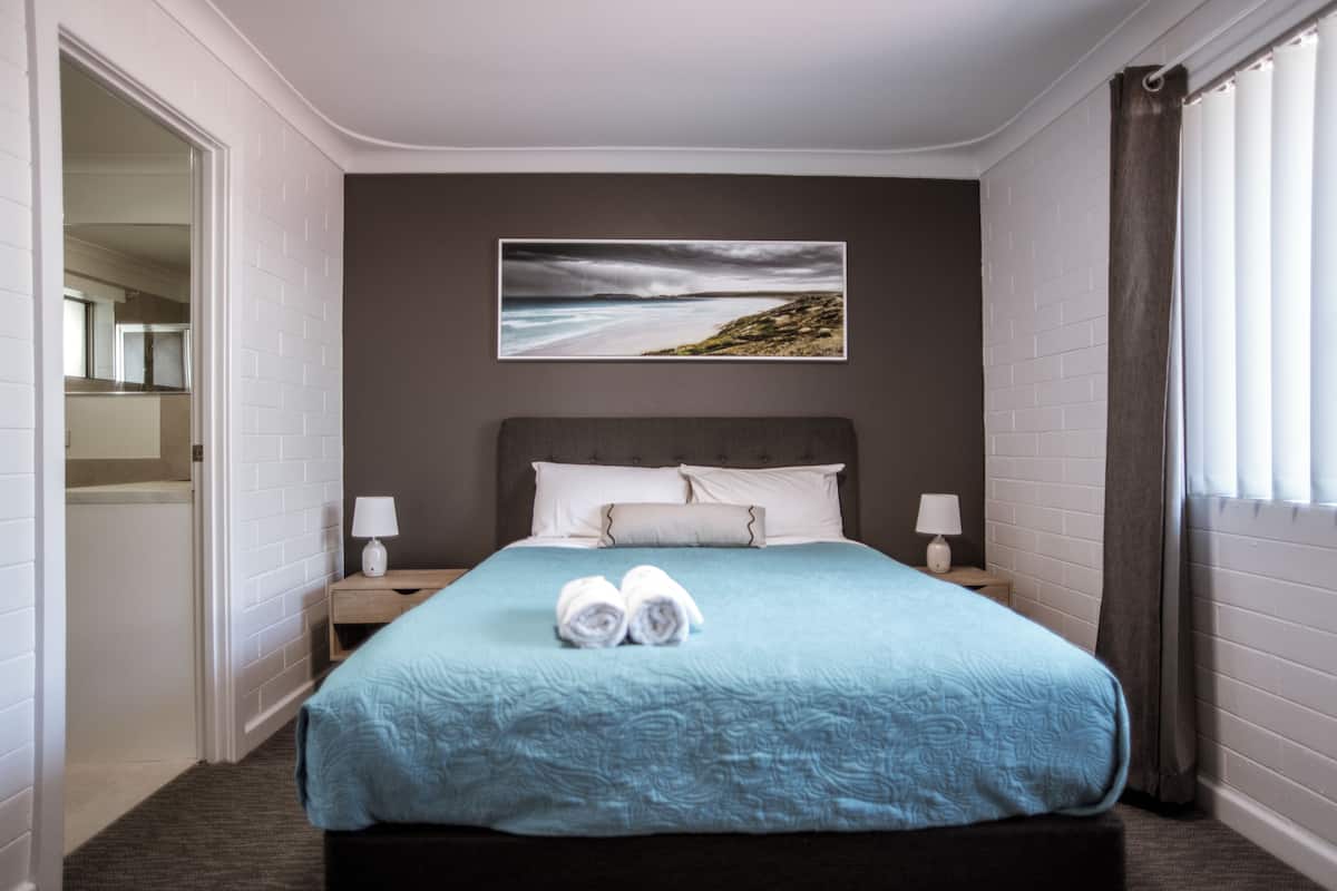 A hotel room with a bed, a painting of a beach, and a window with curtains.