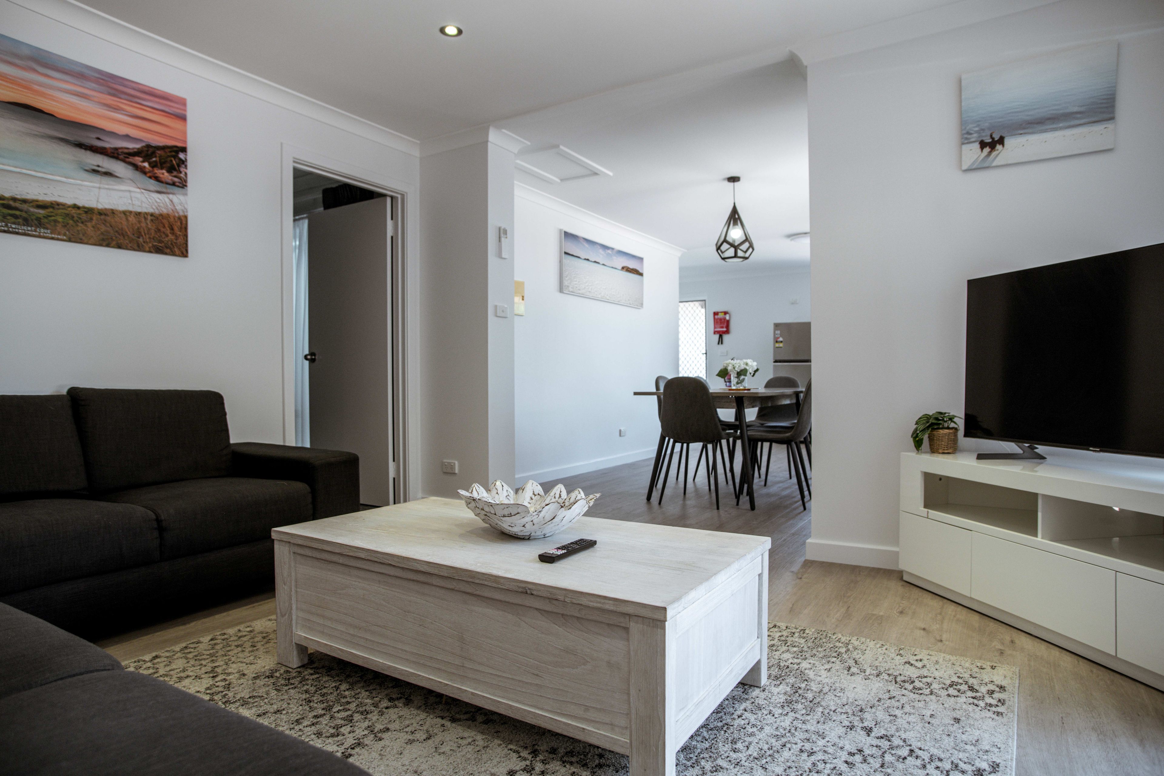 A modern living room with a sofa, coffee table, and TV. There is a dining area with chairs and a table in the background.