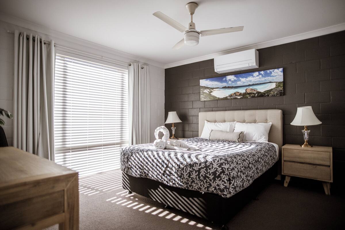 A bedroom with a large bed, a ceiling fan, a nightstand, a lamp, and a painting on the wall.