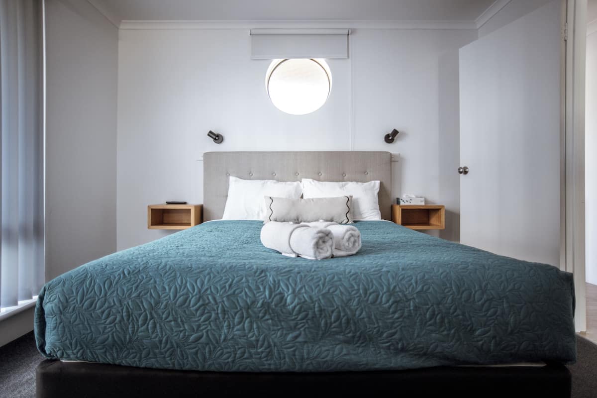 A neatly made bed with a teal quilt, two bedside tables with lamps, and a round window above the bed.