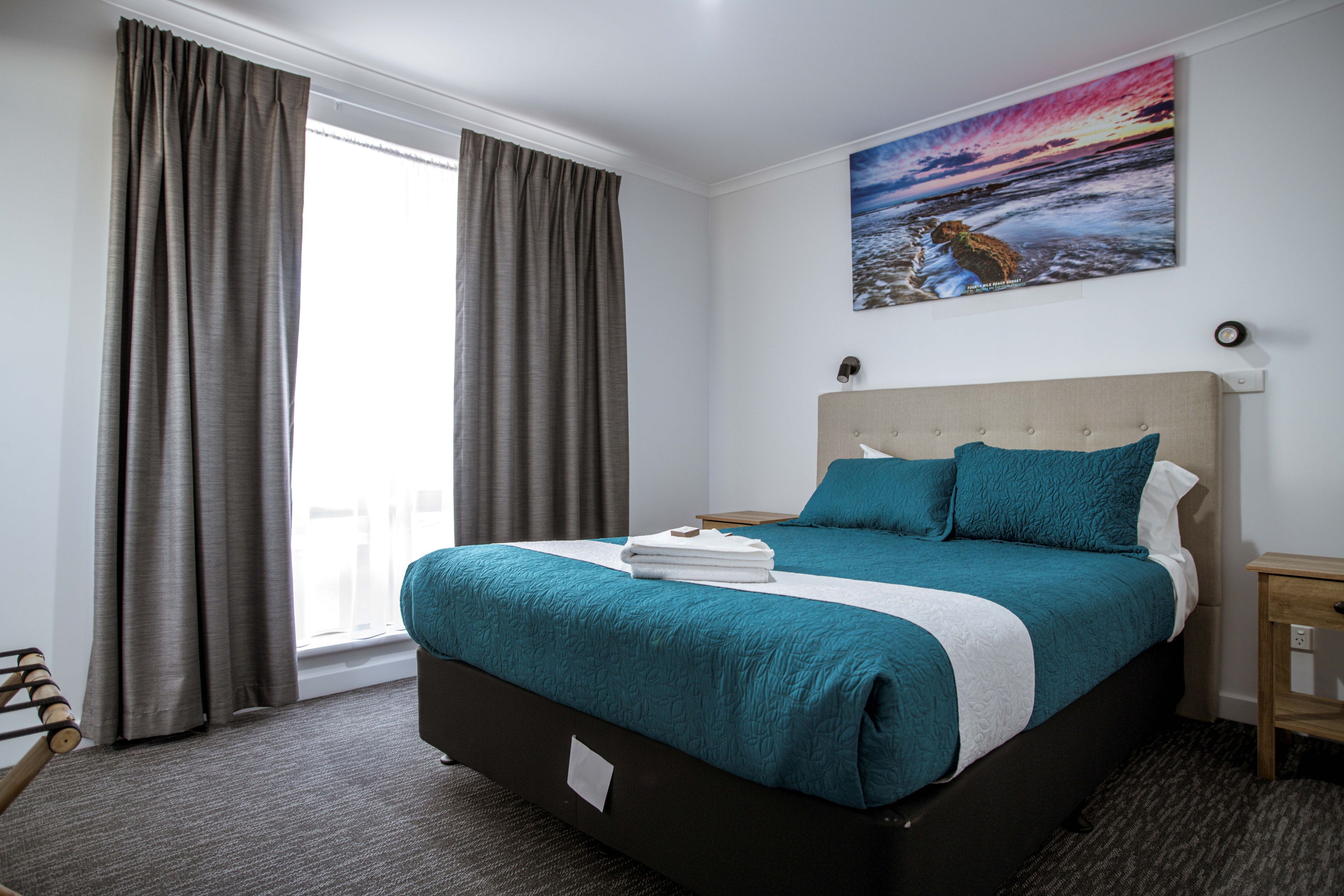 A hotel room with a large bed, a view of the sea through the window, and a painting of a sunset on the wall.
