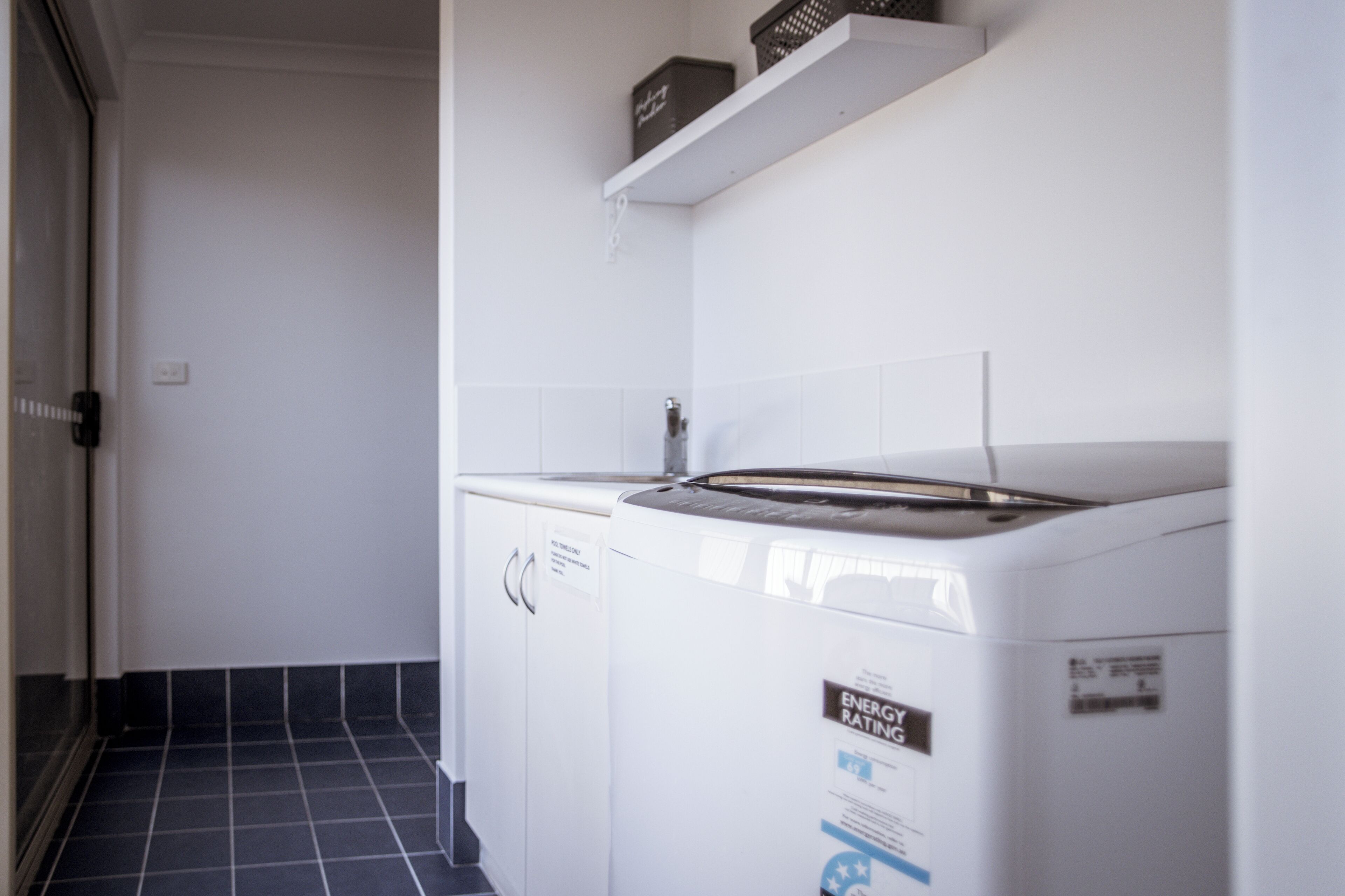 A laundry area with a front-loading washer and dryer, a white shelf above the dryer, and a tiled floor.
