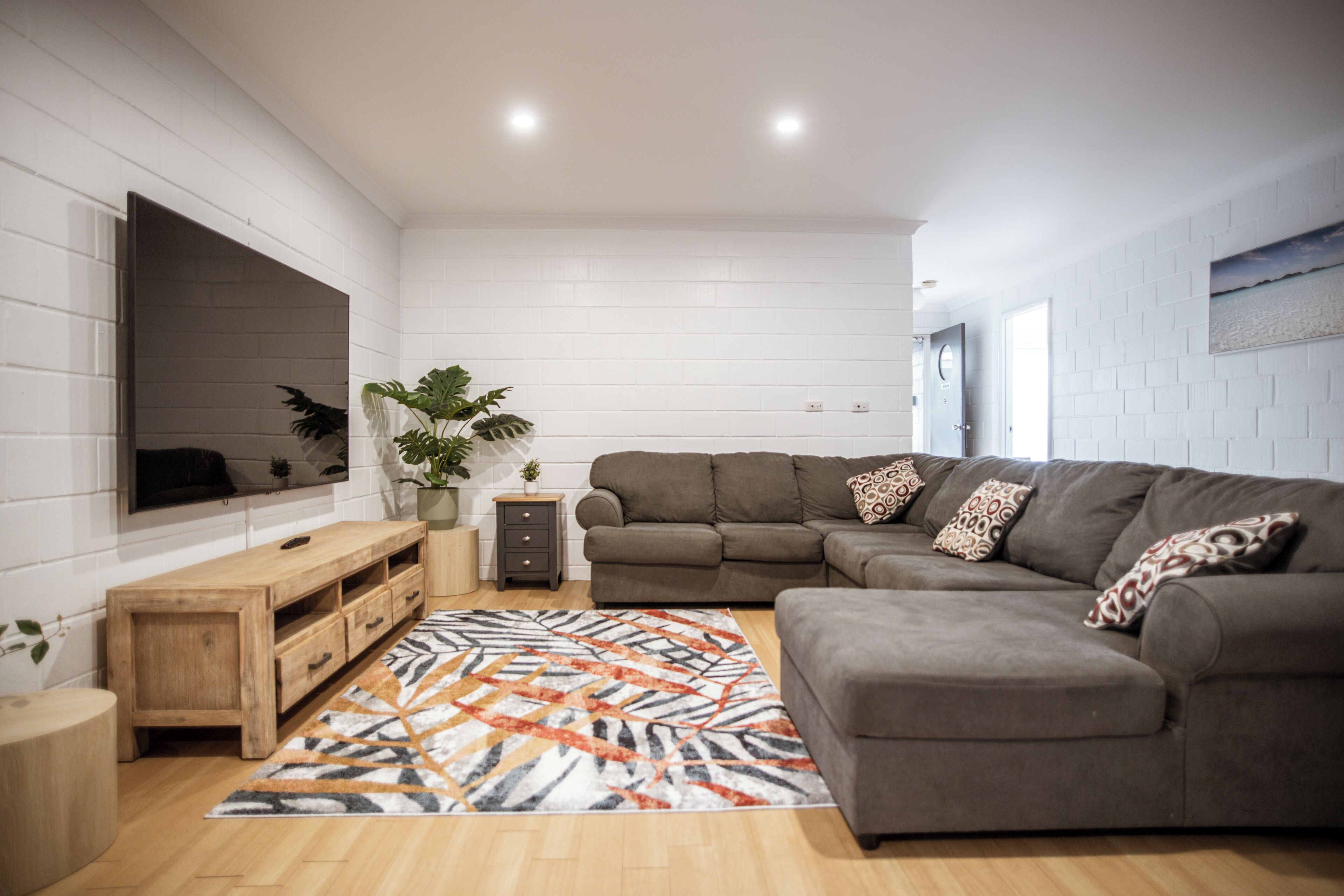 A modern living room with a sectional sofa, a flat-screen TV mounted on the wall, a wooden sideboard, and a patterned rug.