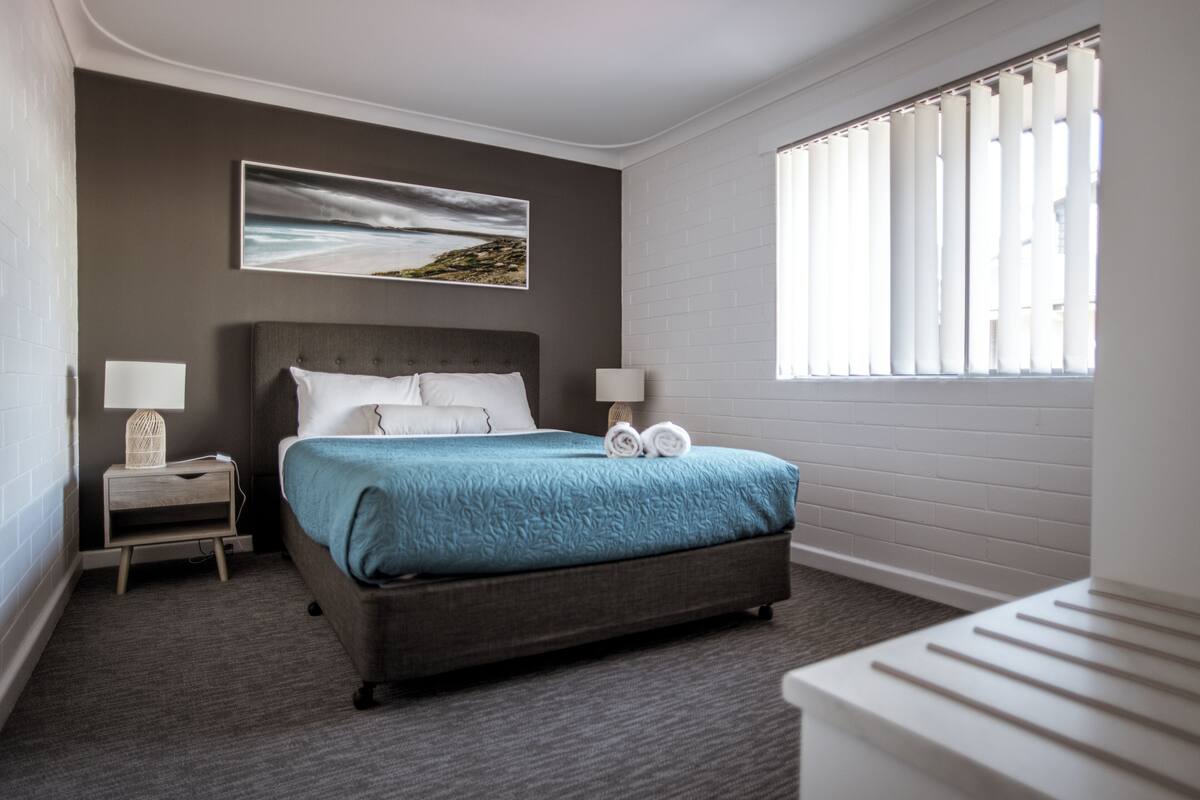 A neatly arranged hotel room with a bed, two bedside tables, a window with blinds, and a painting on the wall.