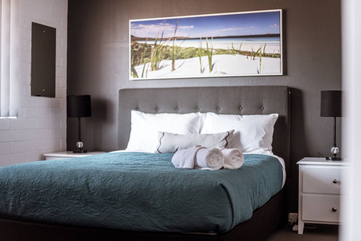 A neatly made bed with a teal bedspread, white pillows, and rolled towels. There are two bedside tables with lamps, and a framed picture of a beach scene hangs above the bed.