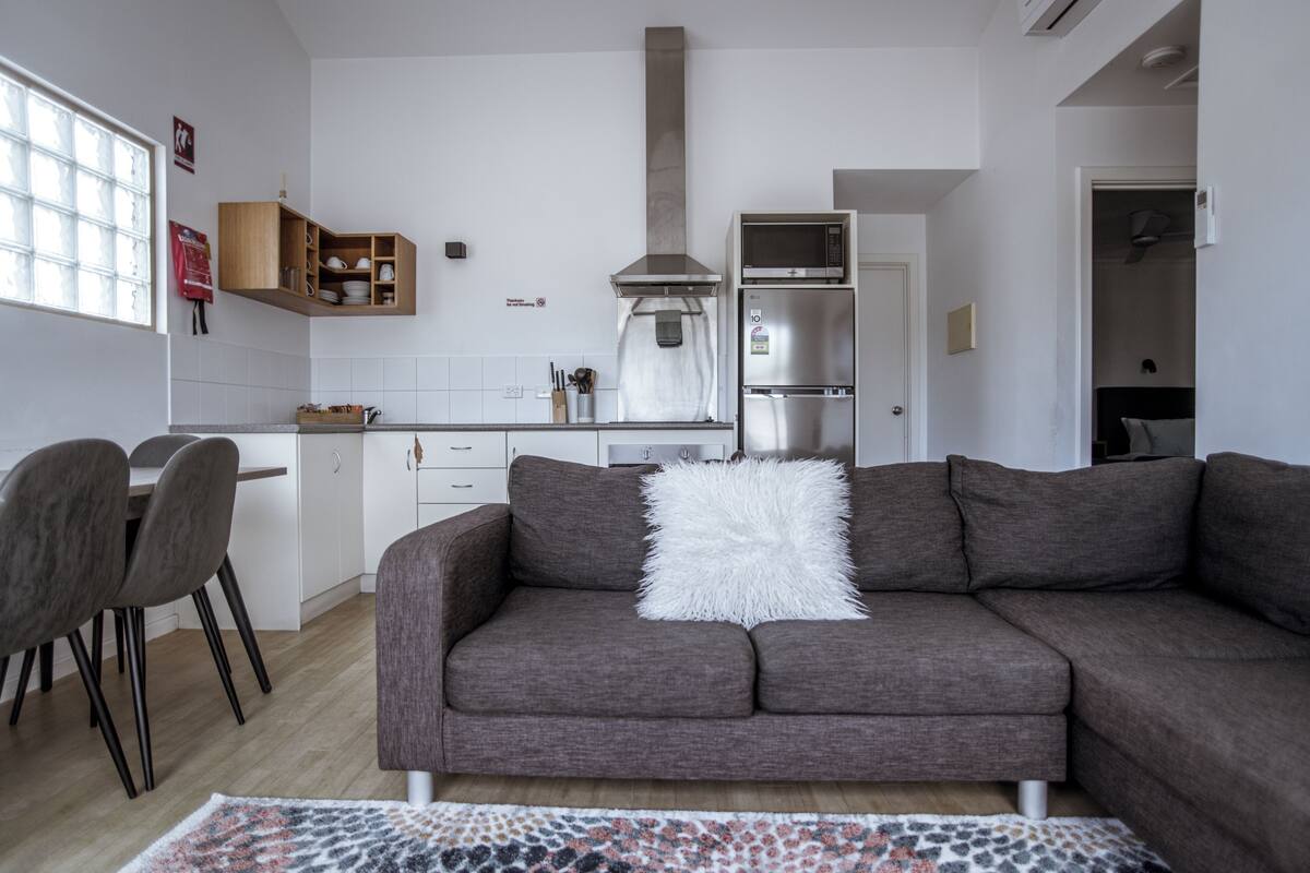 A modern kitchen with a built-in microwave, oven, and a stainless steel refrigerator. There is a dining area with a table and chairs, and a living room with a grey sofa and a white fluffy pillow.