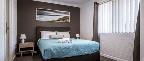 A neatly arranged bedroom with a large bed, a bedside table with a lamp, a wall-mounted picture, and a window with blinds.