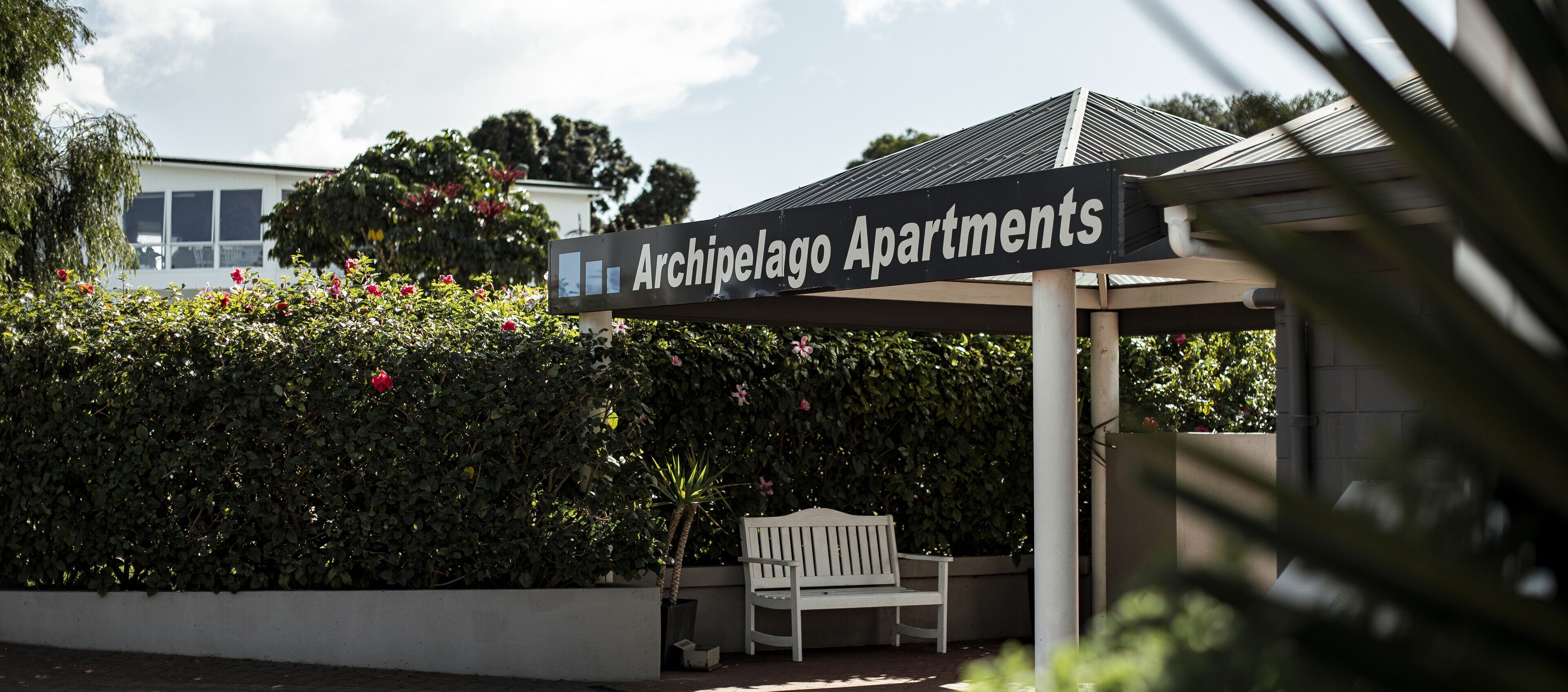 A sign for Archipelago Apartments with a white bench and greenery in the background.
