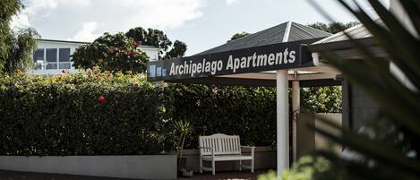 A sign for Archipelago Apartments with a white bench and greenery in the background.