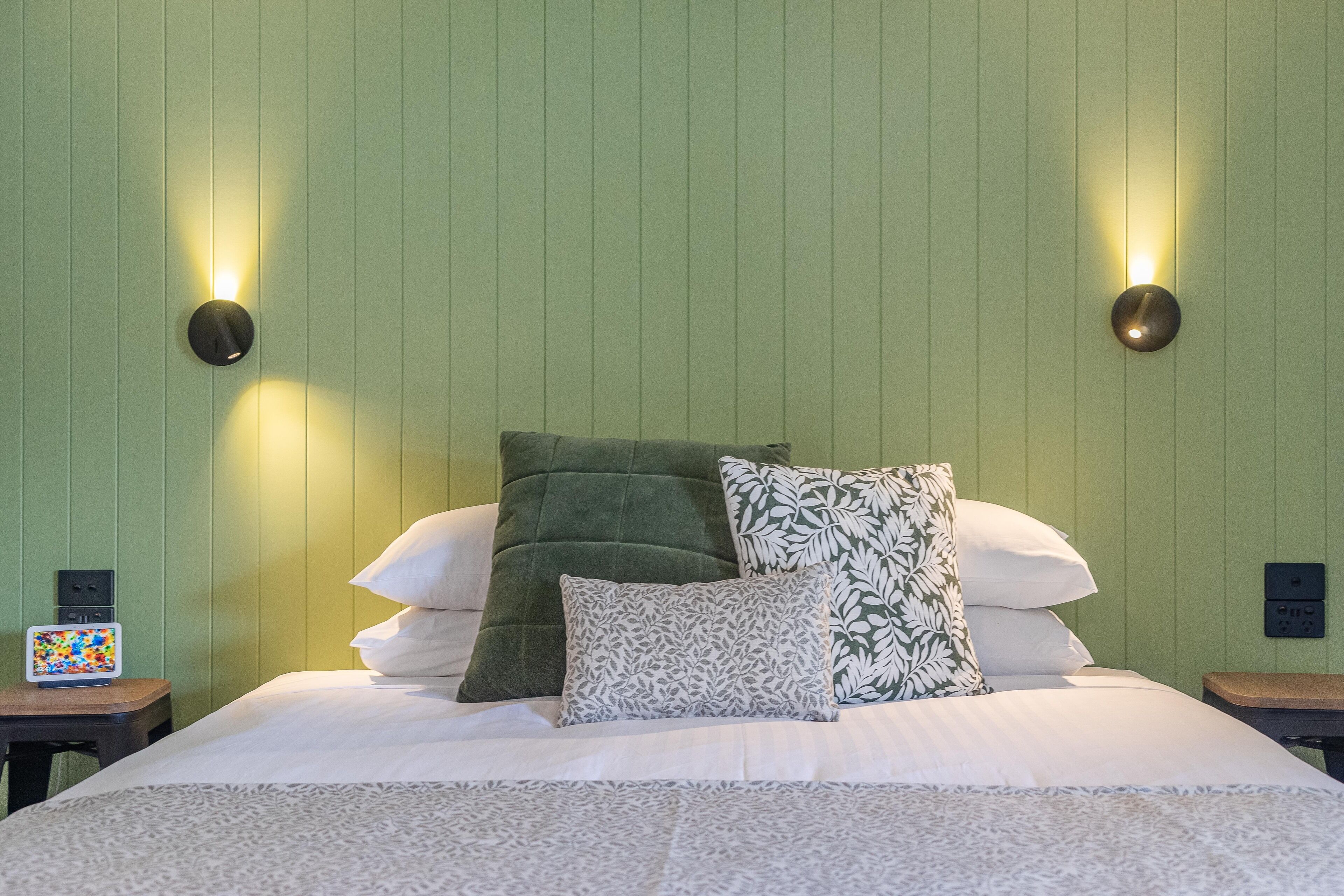 A neatly made bed with patterned bedding and pillows, two wall-mounted light fixtures, and a small table with a colorful item on it.