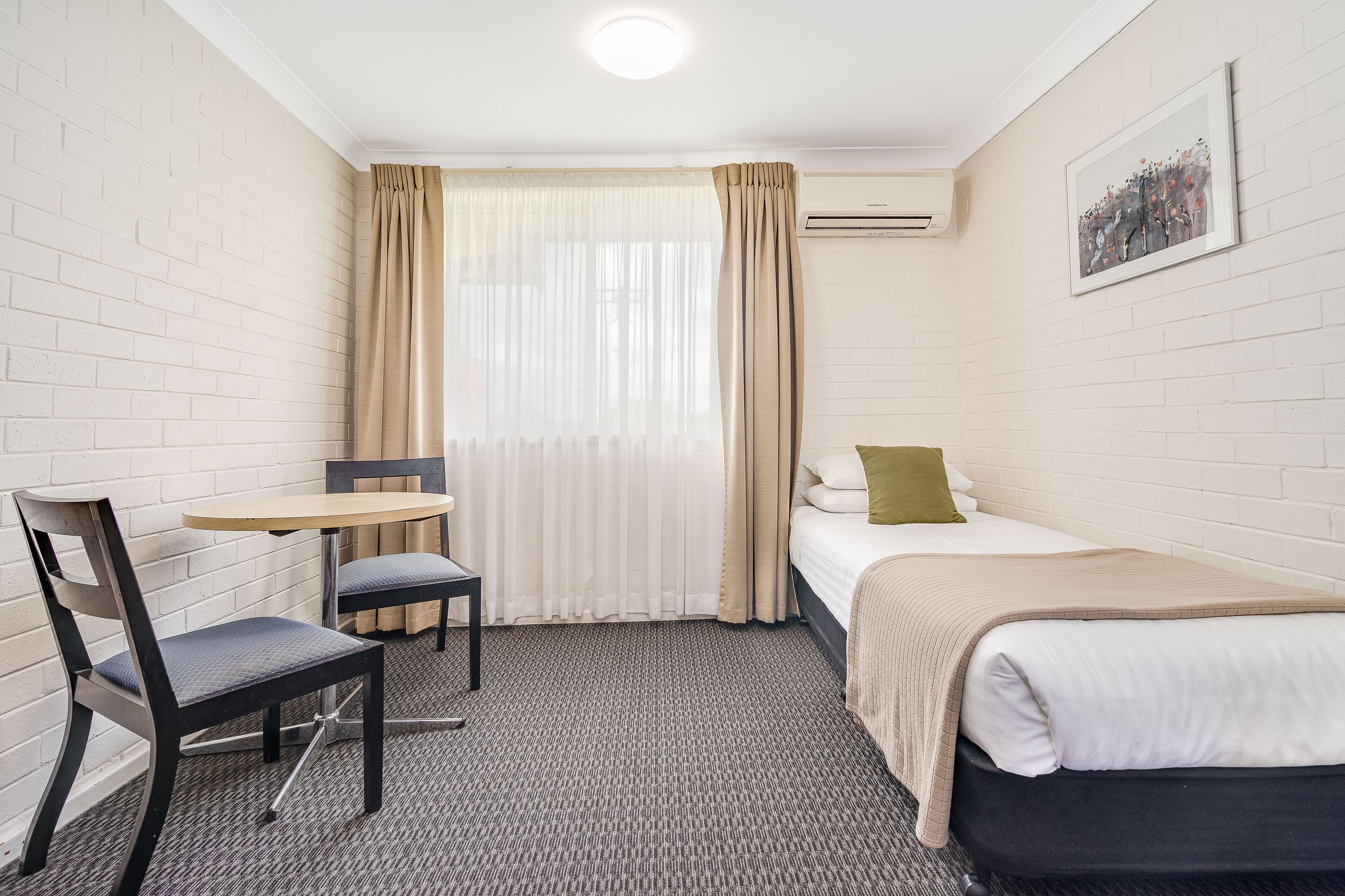 Una habitación de hotel pequeña y ordenada con una cama individual, una silla, una mesa y un ventanal con cortinas.