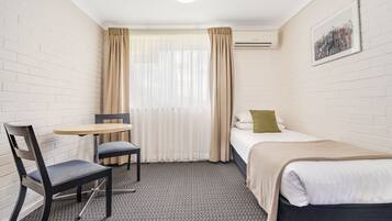 Una habitación de hotel pequeña y ordenada con una cama individual, una silla, una mesa y un ventanal con cortinas.