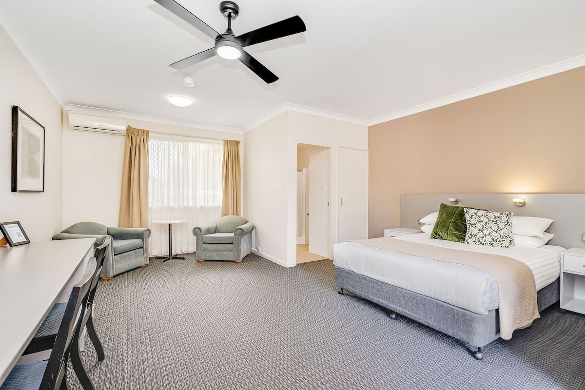 Habitación de hotel con cama, escritorio, dos sillas, ventilador de techo y ventana con cortinas.