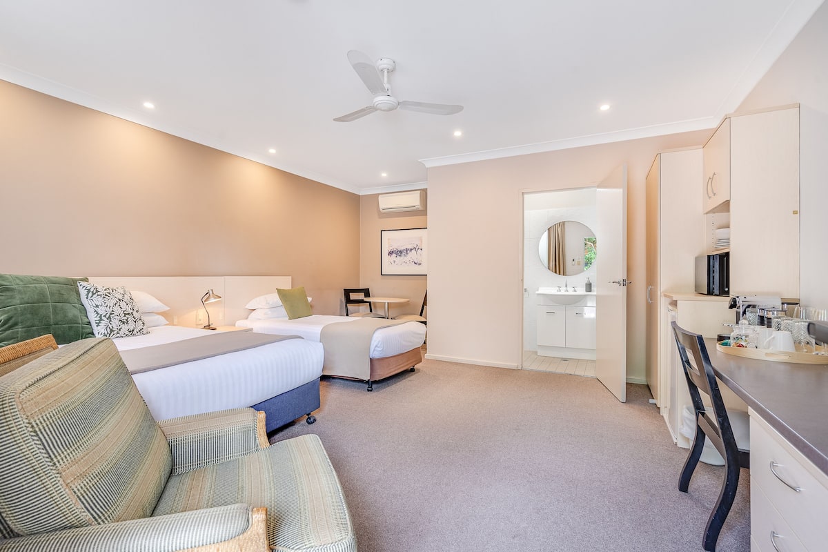 Habitación de hotel con dos camas, un escritorio, una silla, un ventilador de techo y la puerta de un baño.