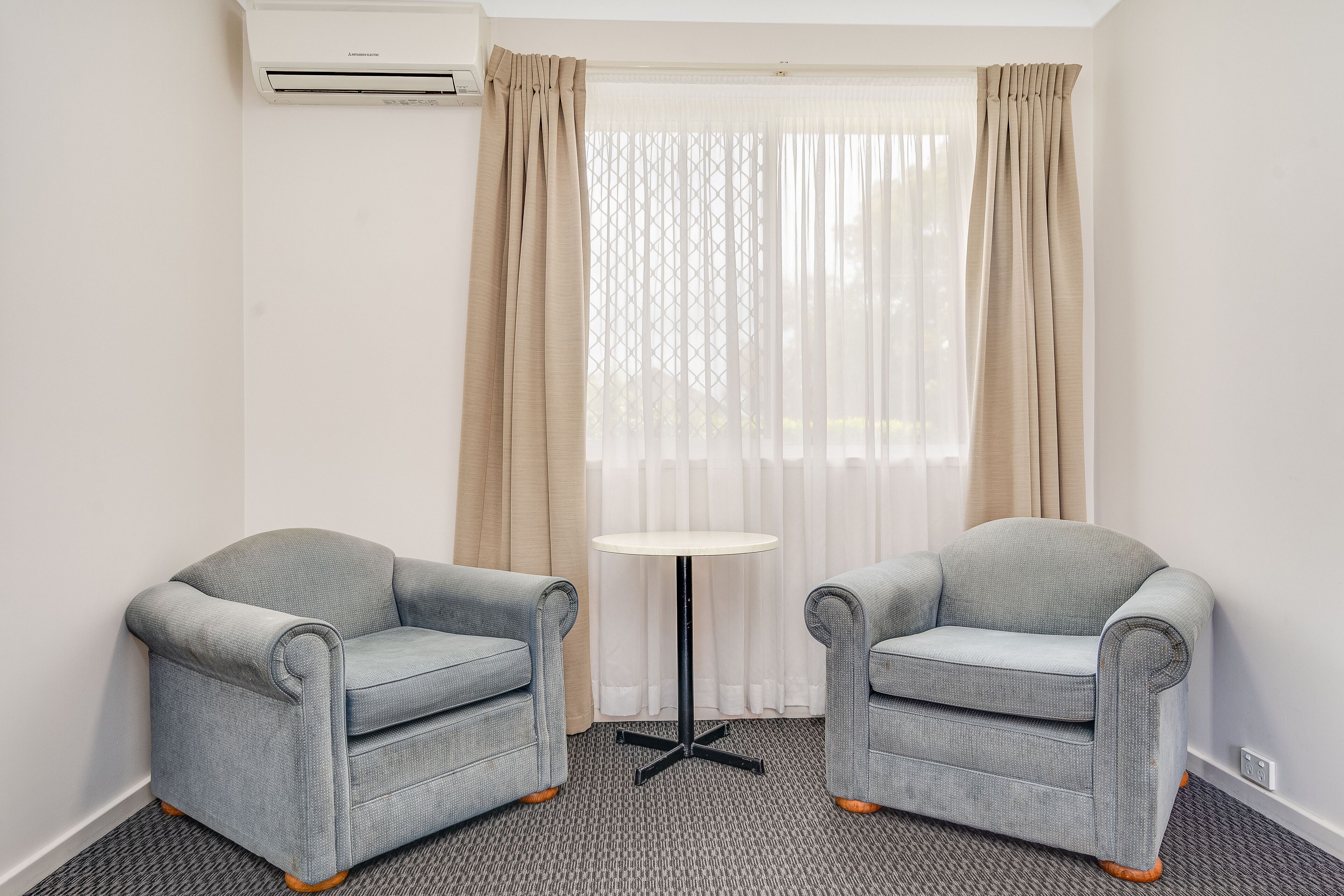 A room with two grey armchairs, a small round table, and a window with sheer curtains.