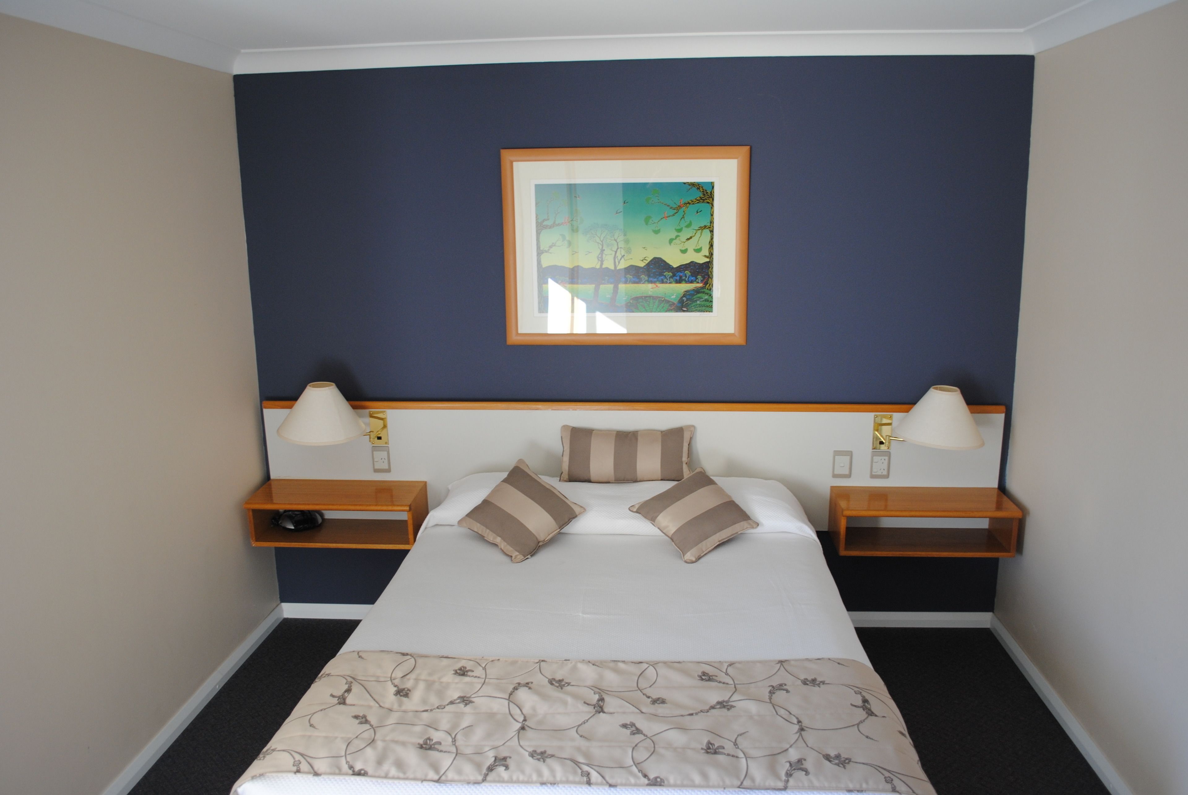 Habitación de hotel con cama, dos mesitas de noche, una lámpara de noche, un cuadro enmarcado en la pared y piso alfombrado.