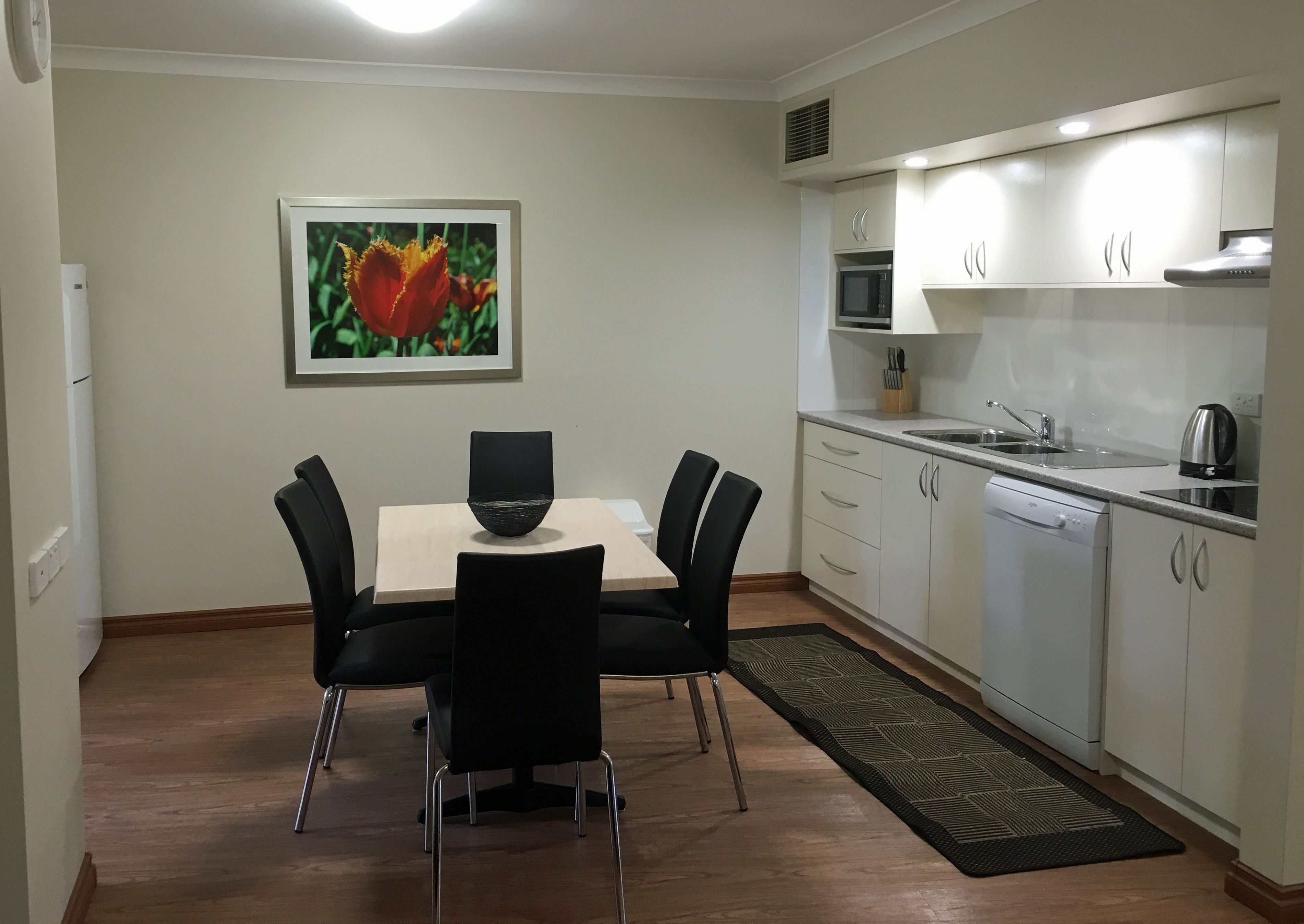 Una cocina compacta con armarios blancos, un microondas y una tetera. Hay una mesa de comedor con cuatro sillas y un cuadro en la pared.