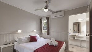 Habitación de hotel con cama, mesita de noche, ventilador de techo, ventana con cortinas y baño con lavabo y espejo.