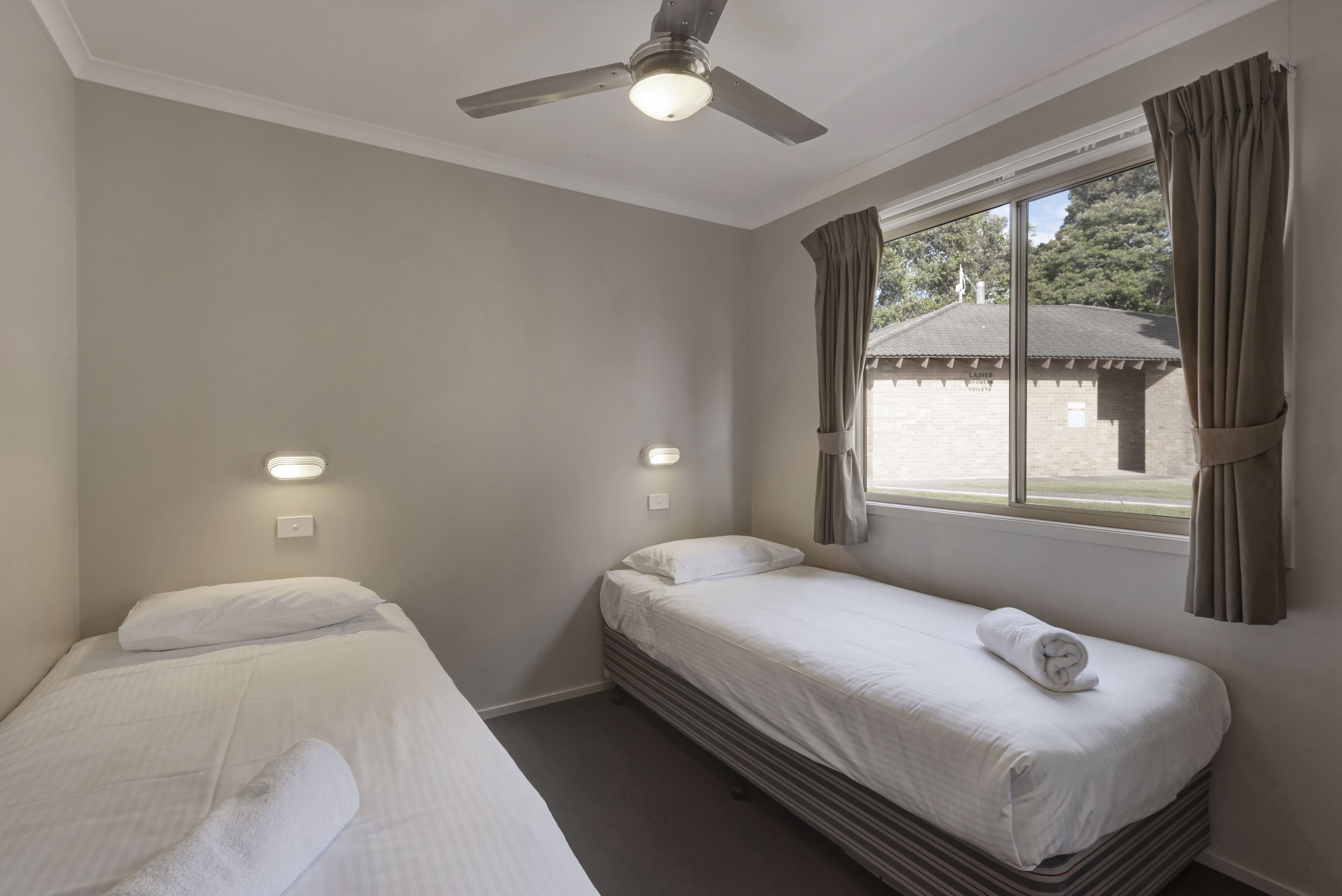 A room with two single beds, a ceiling fan, a window with curtains, and a view of a building outside.