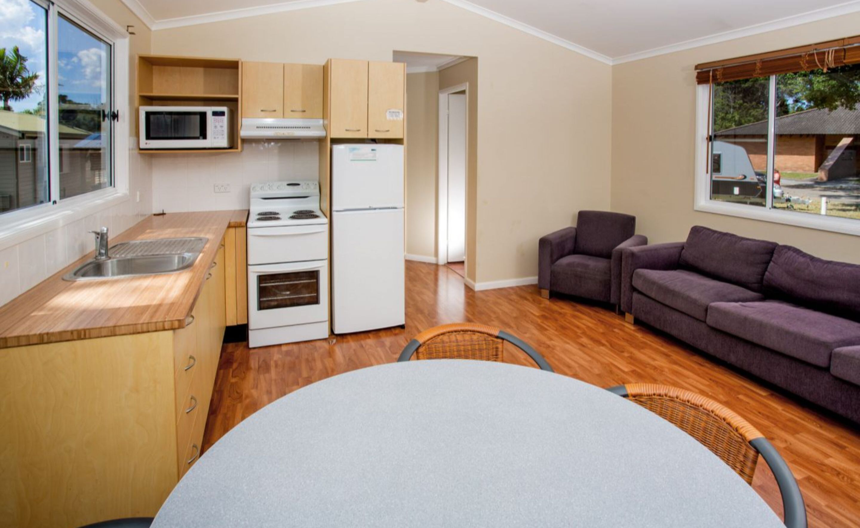 A compact kitchen with a microwave, stove, and refrigerator, a dining table with chairs, and a sofa in a living area with a window.