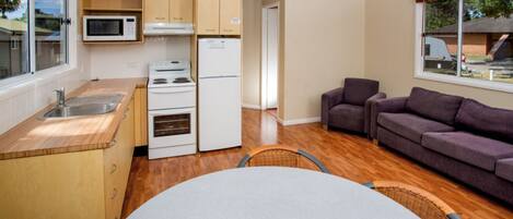 A compact kitchen with a microwave, stove, and refrigerator, a dining table with chairs, and a sofa in a living area with a window.