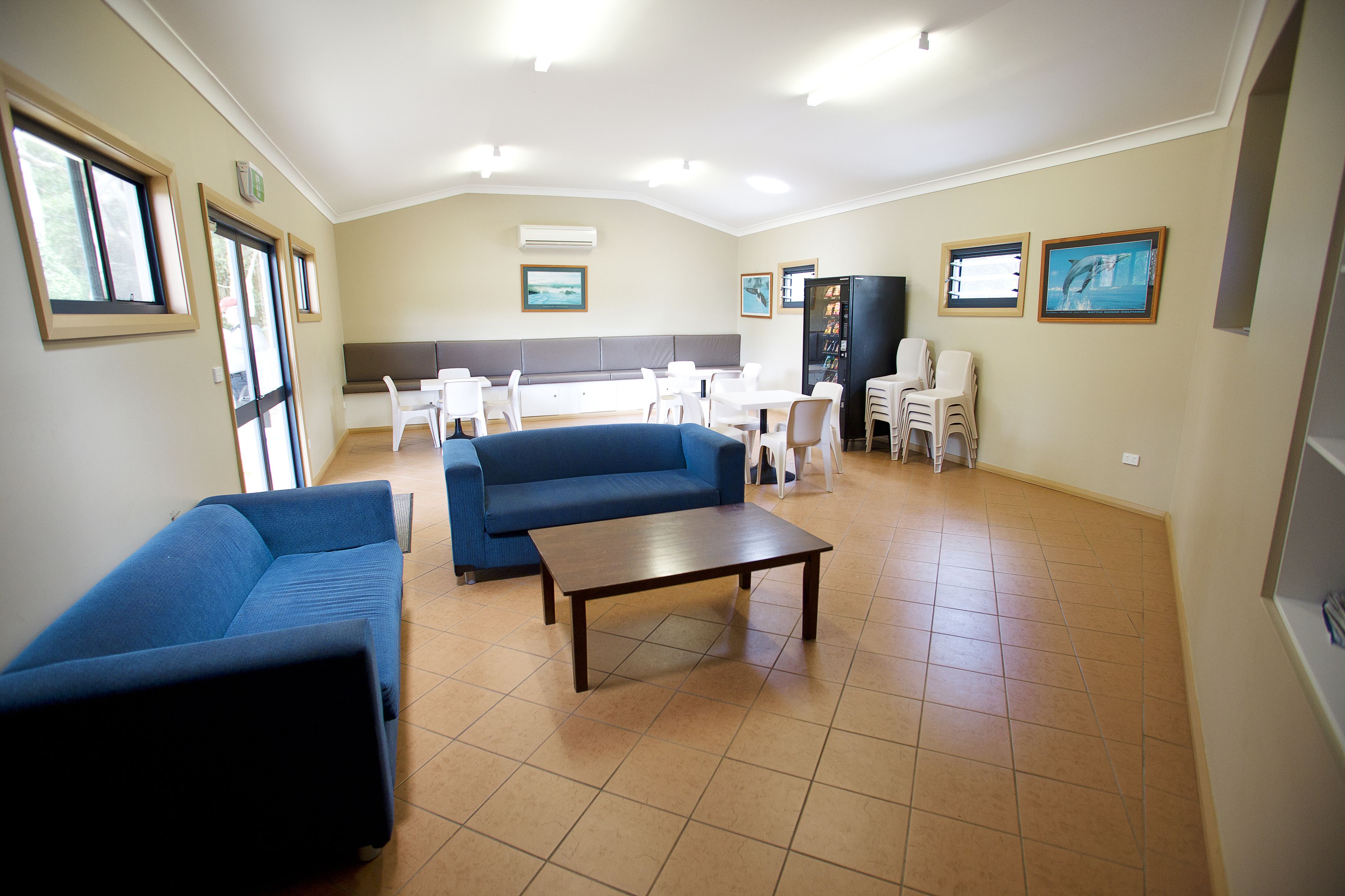 A spacious living area with a blue sofa, a wooden coffee table, and a dining table with chairs. There are framed pictures on the walls and a black cabinet.
