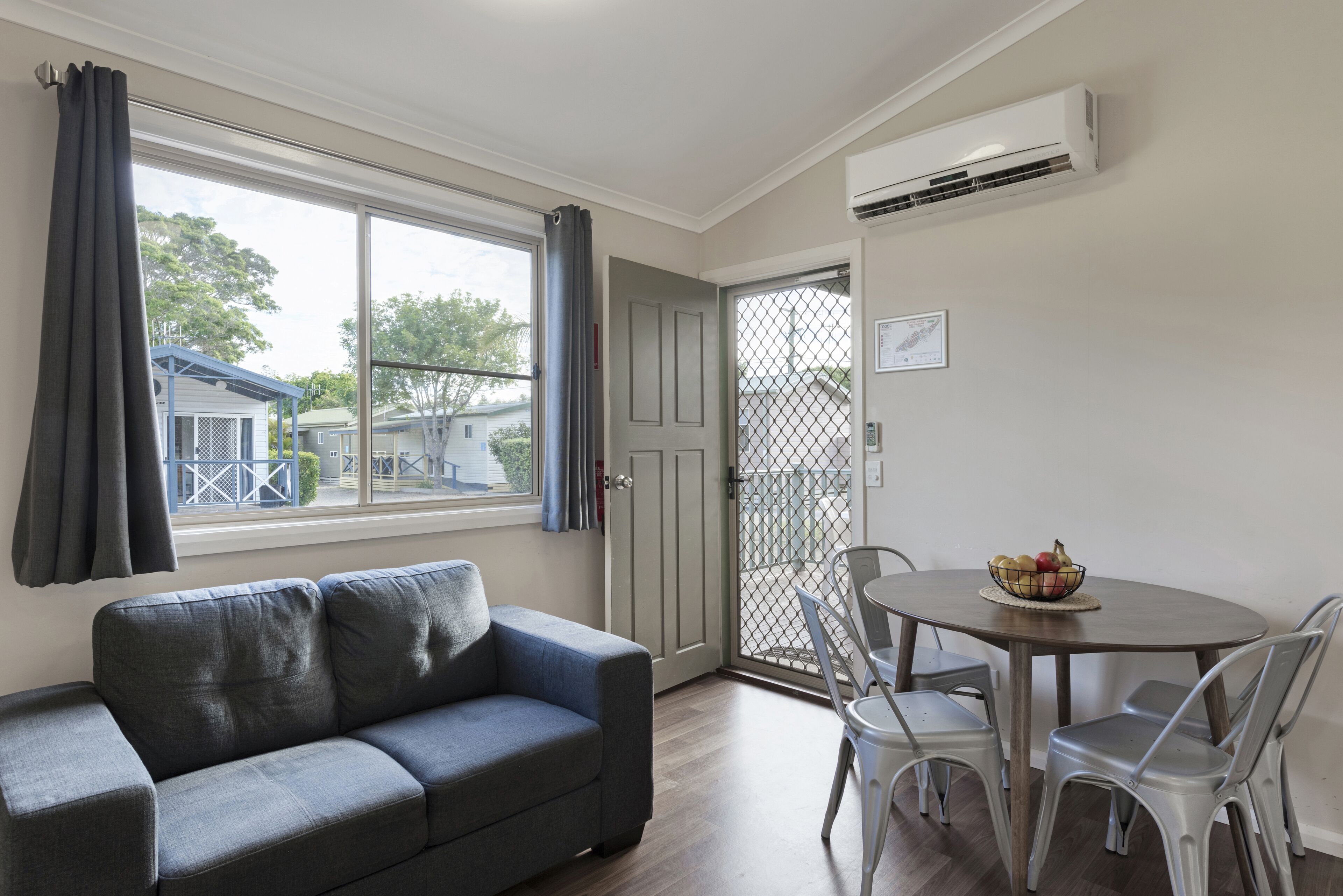 A room with a grey sofa, a dining table with chairs, a window with curtains, and an air conditioning unit.
