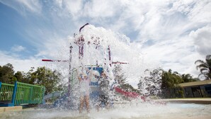 Una fuente de agua con múltiples chorros, rodeada de una zona pavimentada y una valla verde.