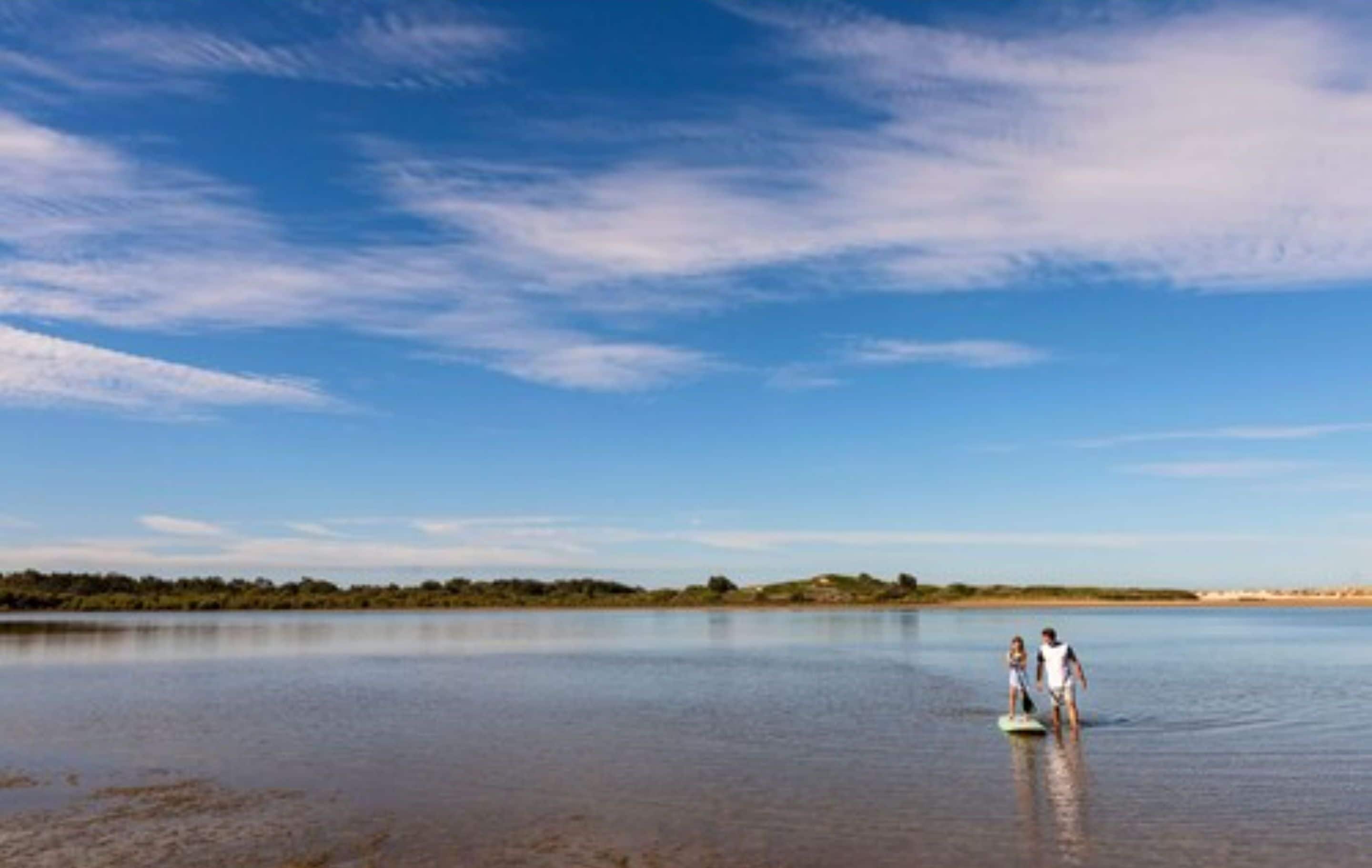 Un lac paisible où se trouvent deux personnes dans l’eau, entouré d’un paysage plat et sous un ciel dégagé.