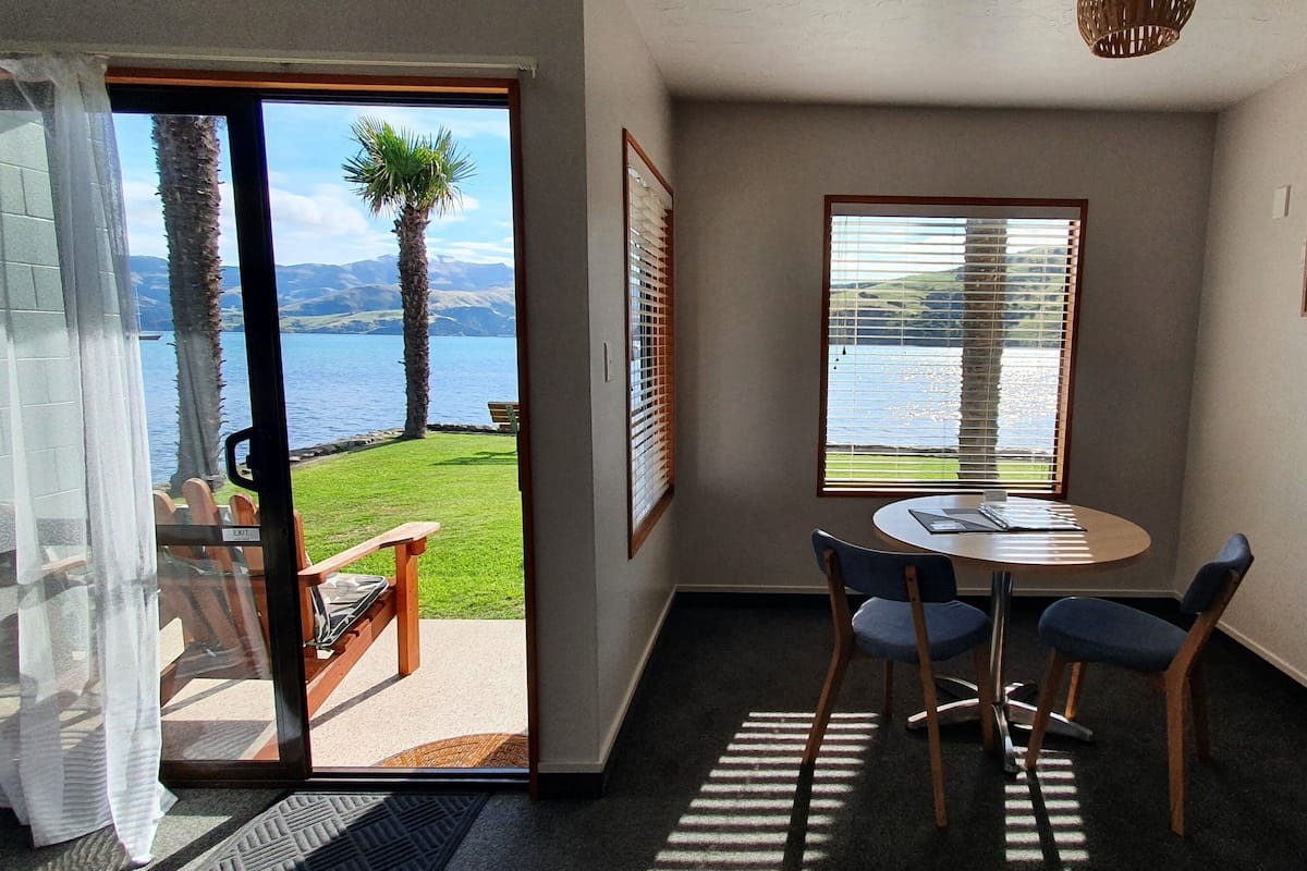 A room with a view of a lake, a round table with chairs, and a door leading to an outdoor area with a bench and chairs.
