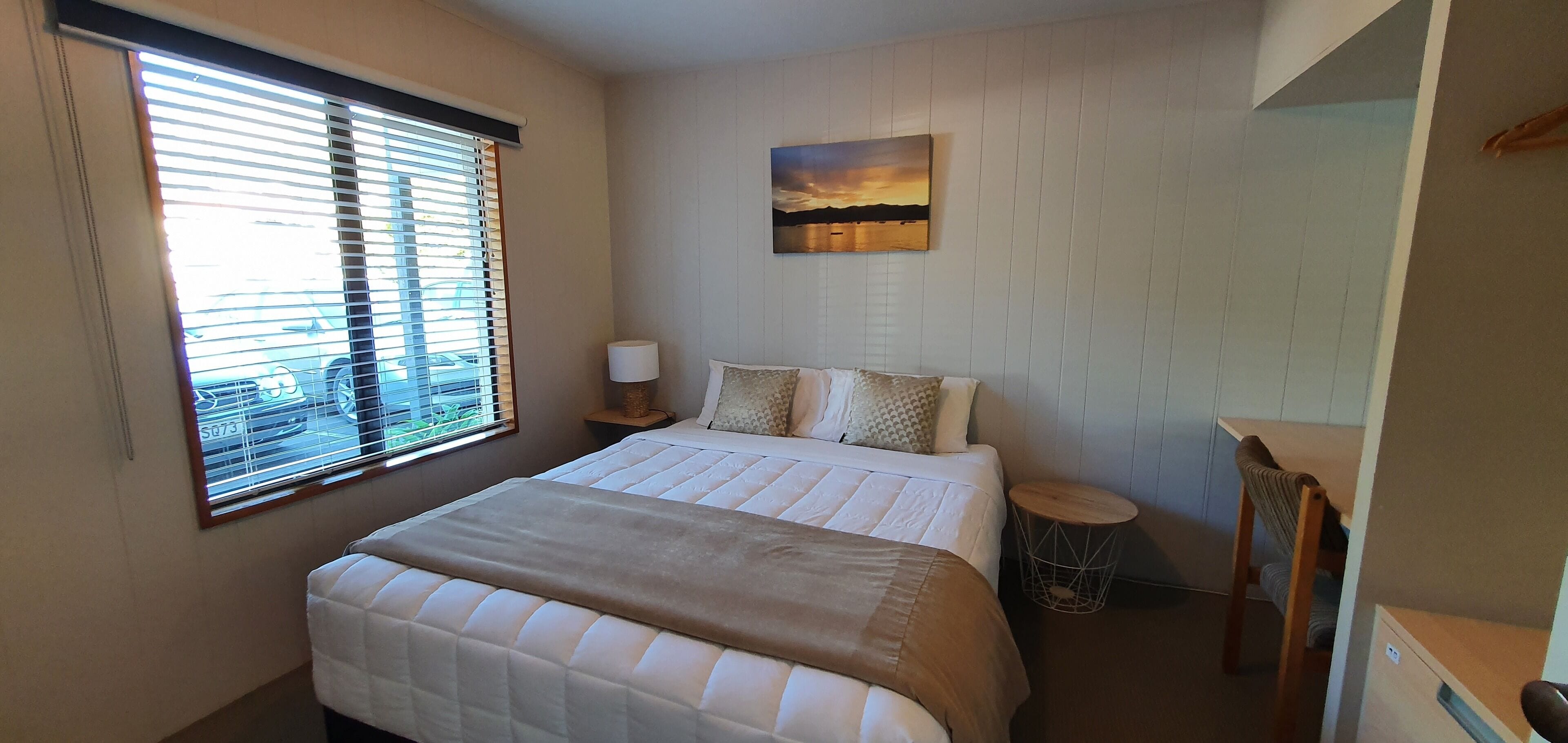 A bedroom with a bed, a bedside table with a lamp, a round side table, a chair, and a painting on the wall.