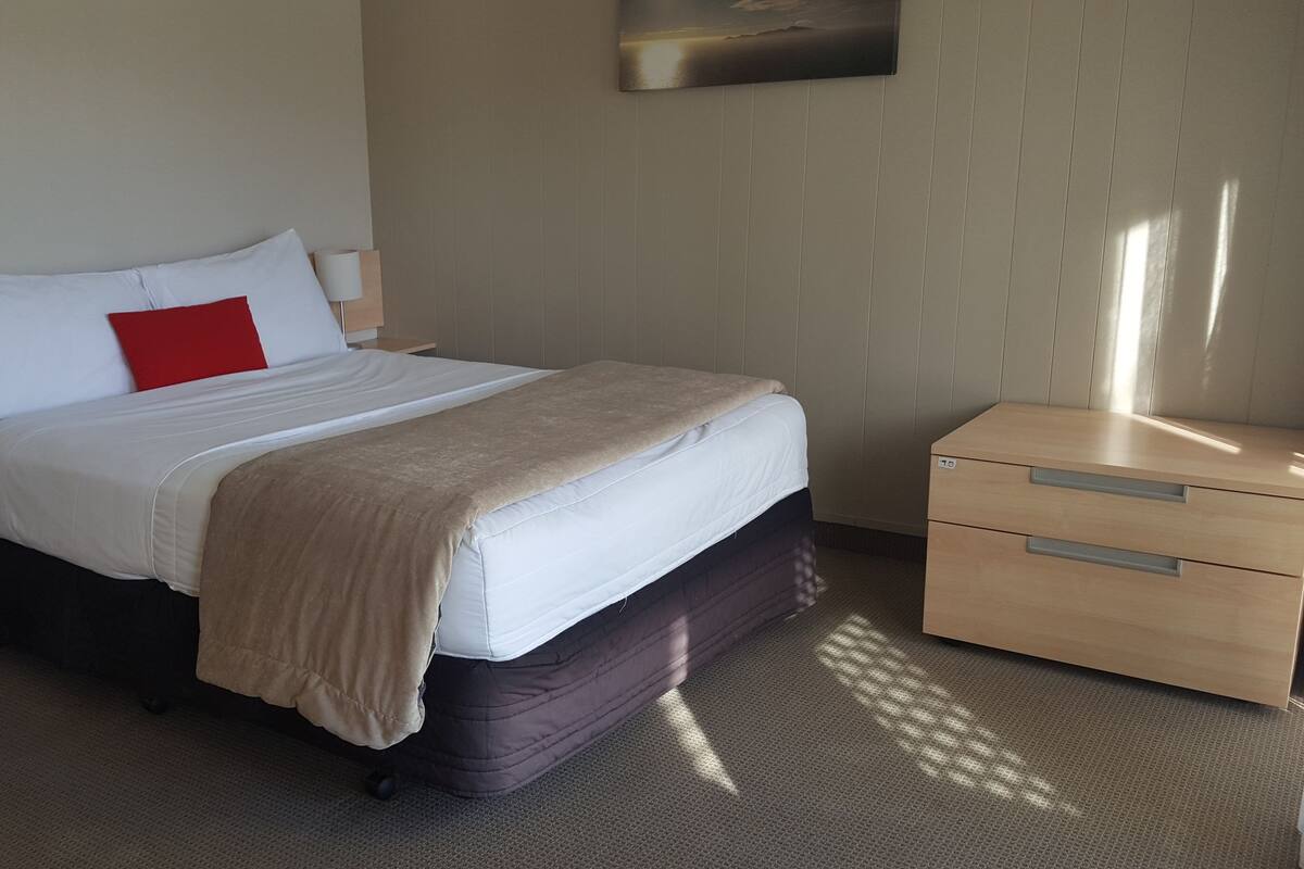 A neatly made bed with white and red pillows, a beige blanket, and a wooden nightstand with drawers.