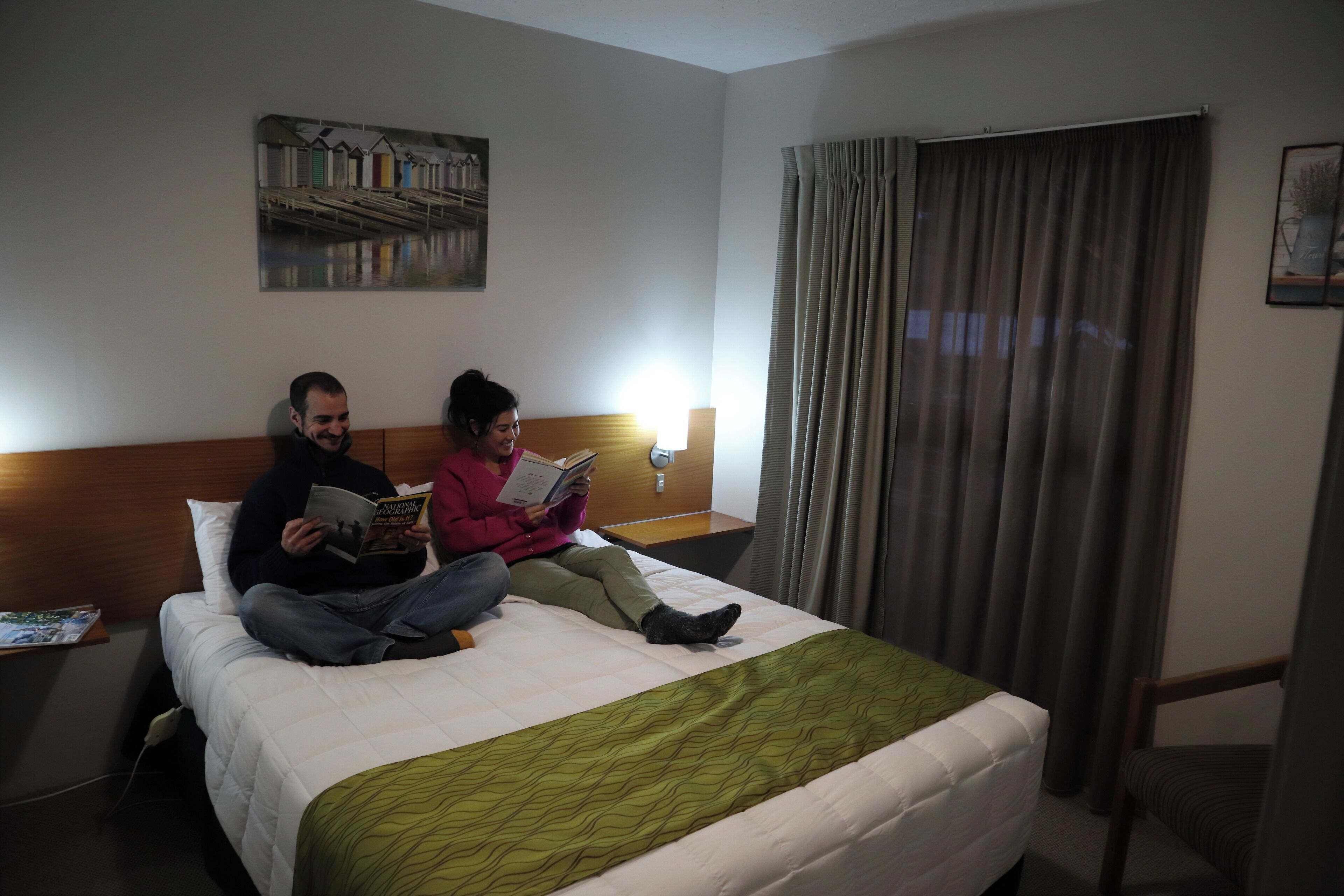 A hotel room with a bed, a nightstand, a lamp, a painting on the wall, and a window with curtains.