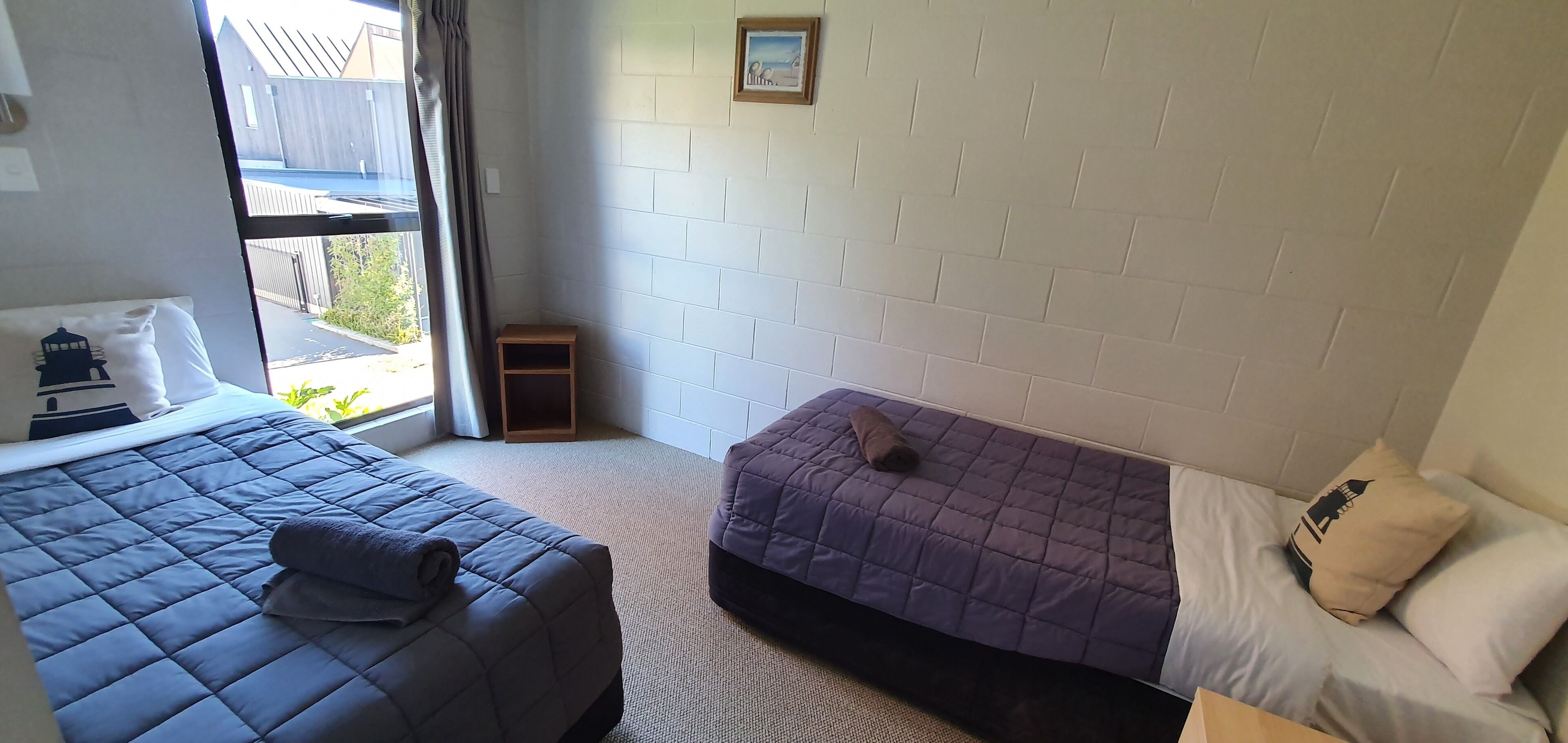 A bedroom with two beds, a window with curtains, a small wooden shelf, and a picture on the wall.