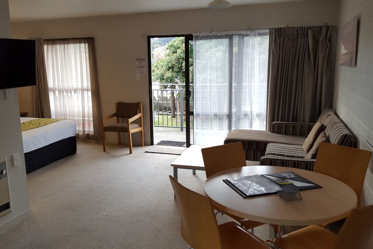 A hotel room with a bed, a TV, a dining table with chairs, a sofa, and a sliding glass door leading to an outdoor area.