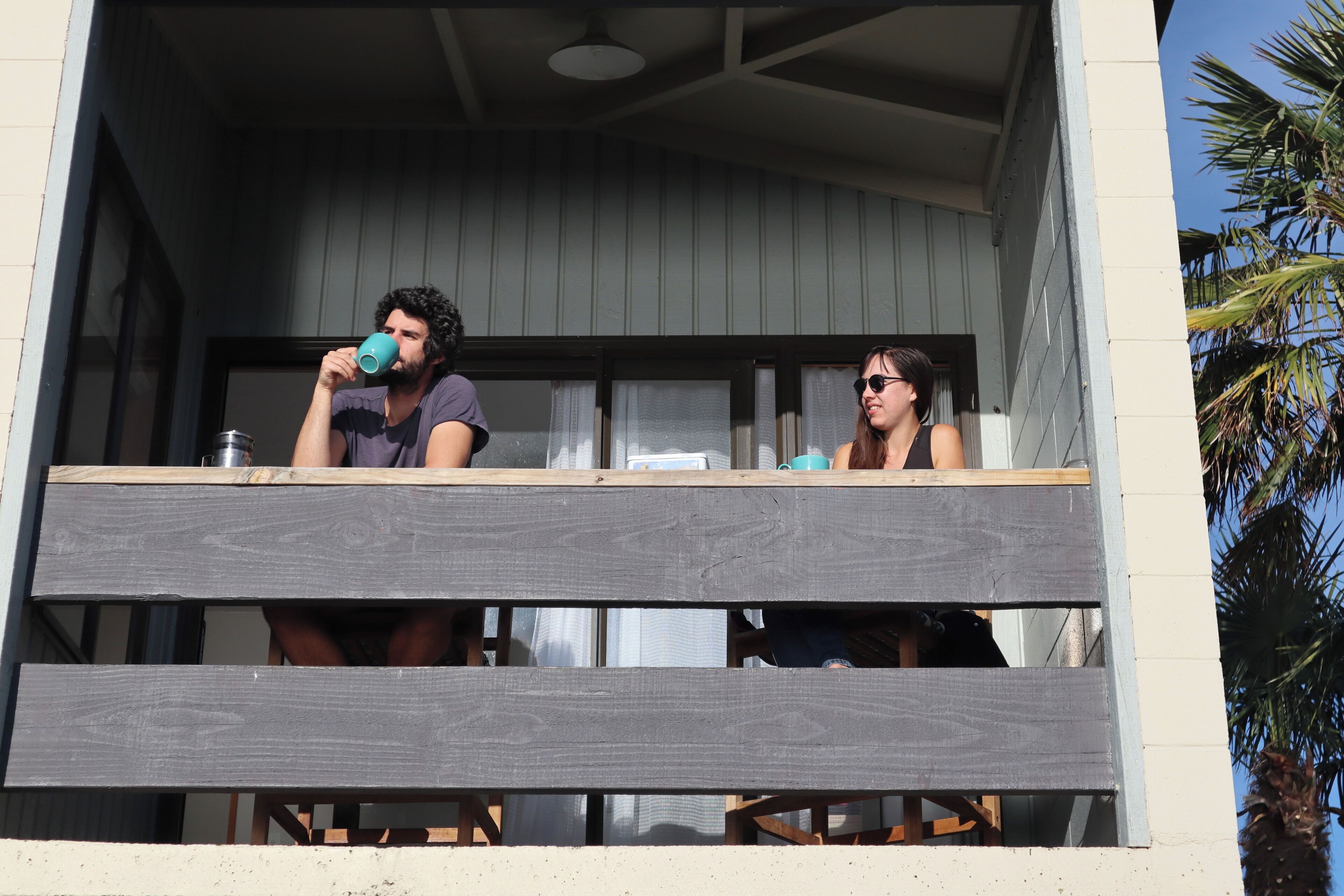 A balcony with wooden railings, two people seated, and a palm tree in the background.
