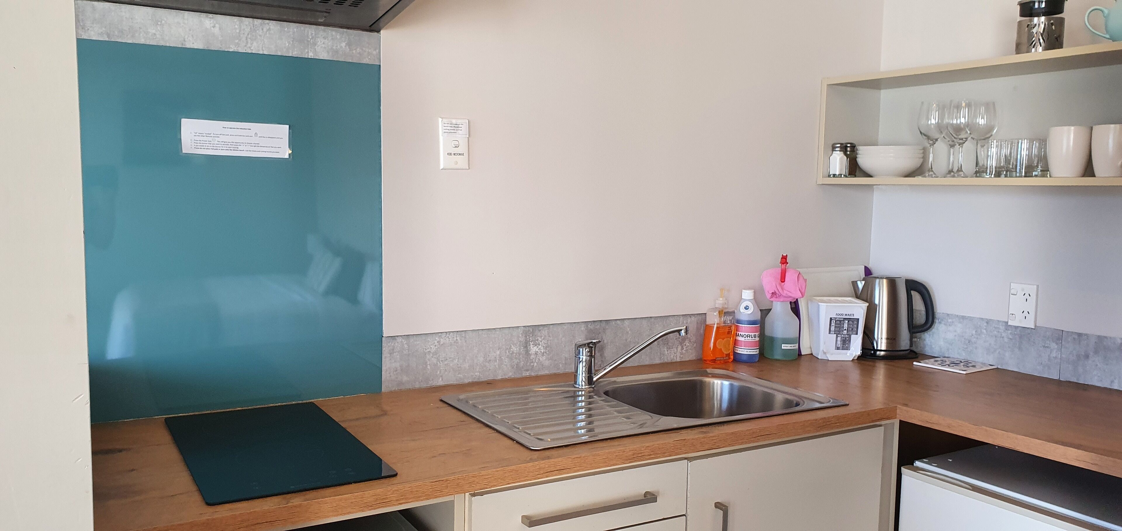 A kitchen with a stainless steel sink, a kettle, and a coffee maker. There are bottles of hand soap and a pink cloth.