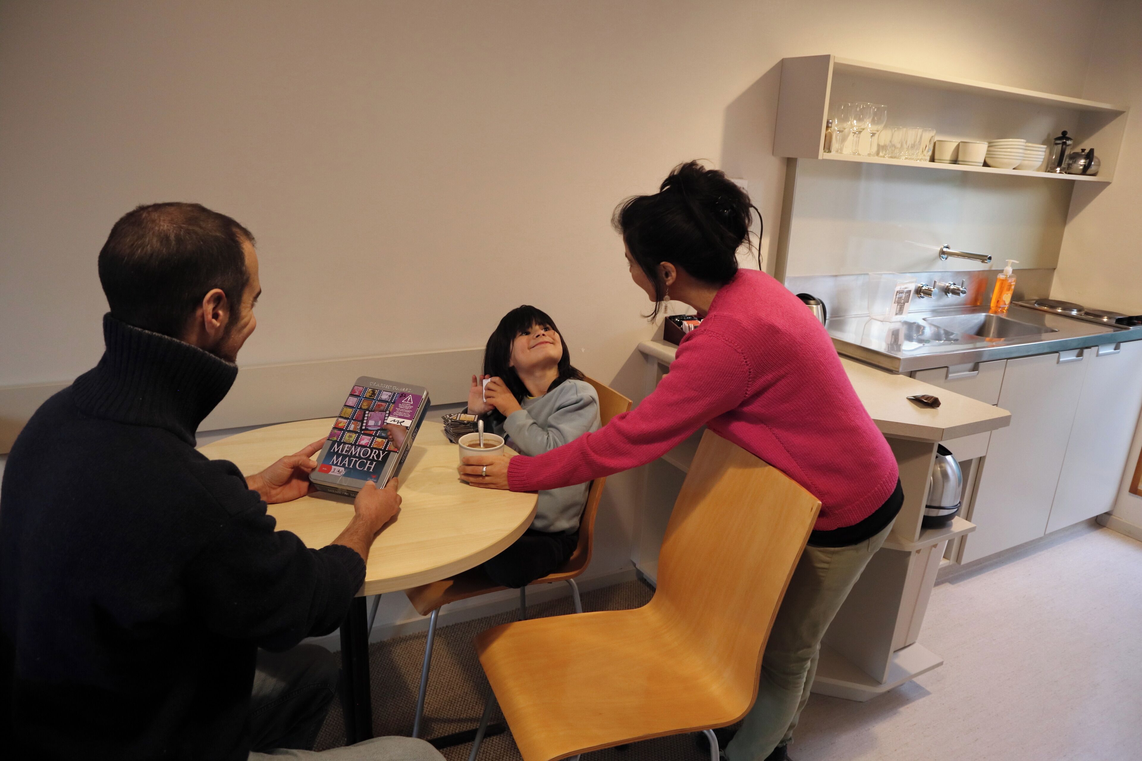 A modern kitchen with a round dining table, a person reading a book, and another person pouring a drink.