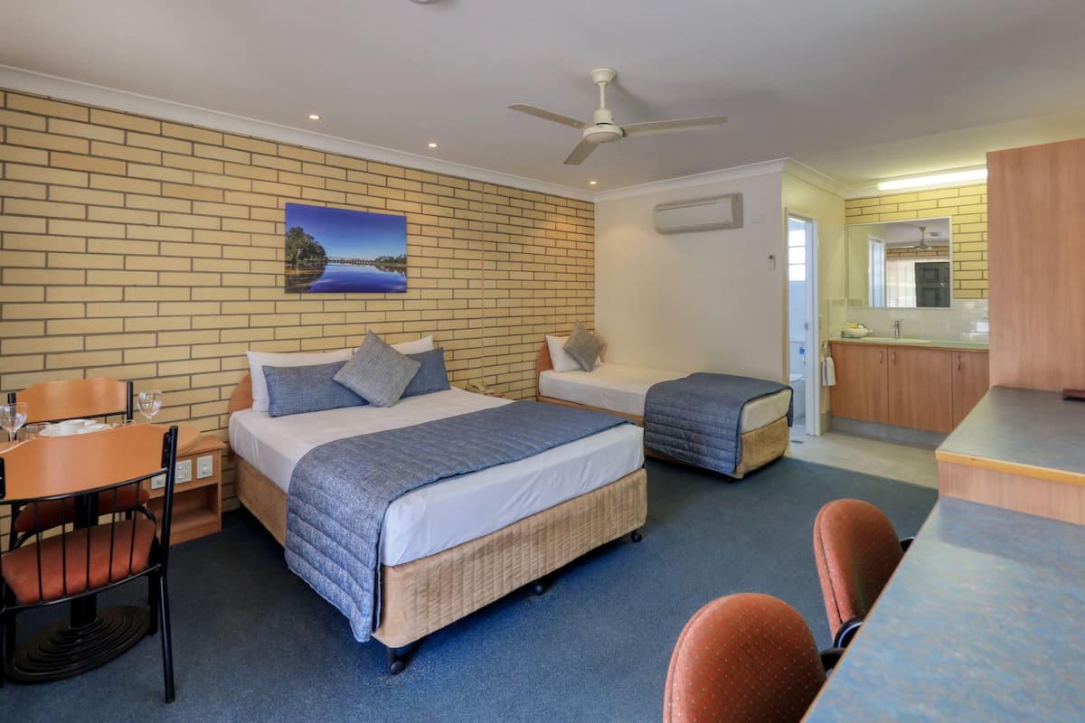 A hotel room with a brick-patterned wall, a bed with a blue blanket, a small desk with a chair, a dining table, and a kitchenette.