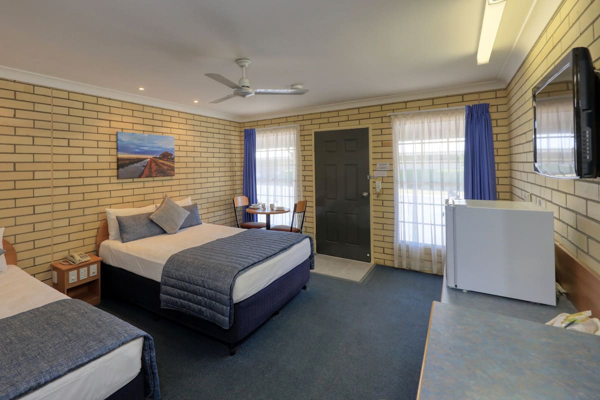 A hotel room with two beds, a desk, a chair, a TV, a ceiling fan, and a brick wall.