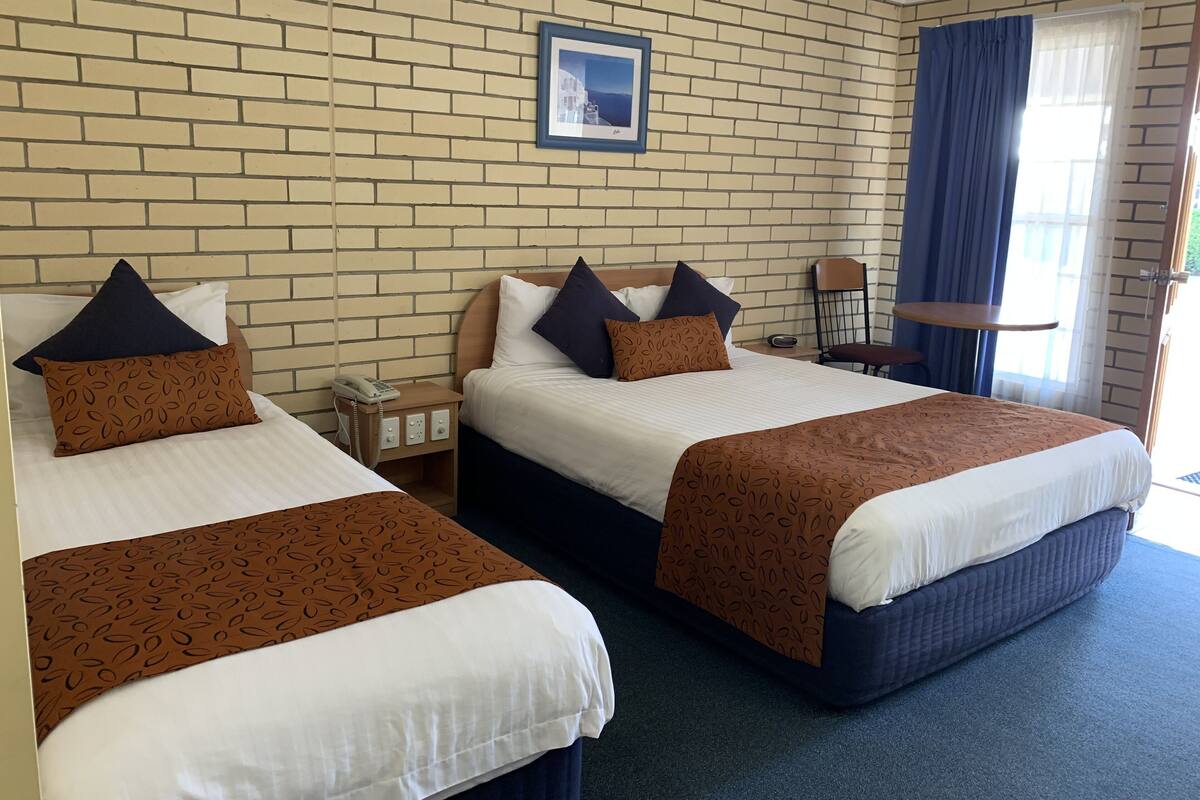 A hotel room with two beds, a brick wall, a framed picture, a nightstand with a phone, and a chair.
