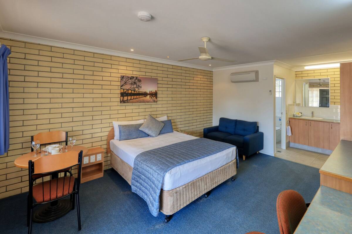 A hotel room with a bed, a desk, a chair, a sofa, a bathroom with a mirror, and a ceiling fan.