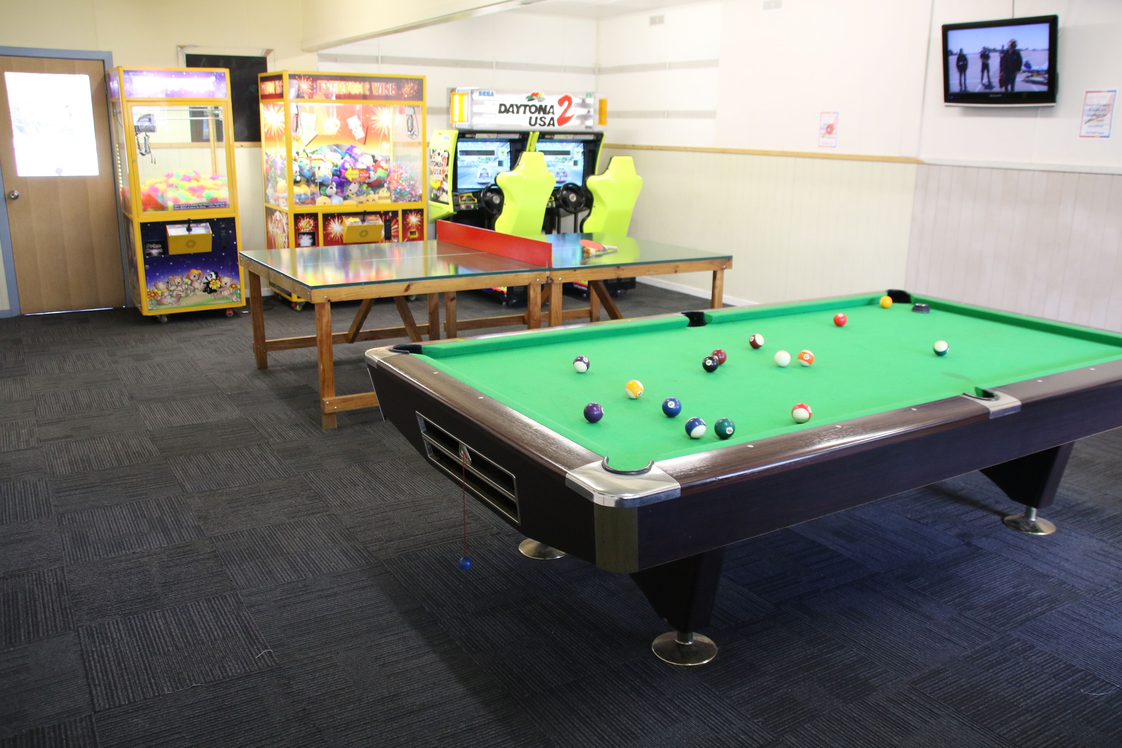 Una mesa de billar con bolas, una mesa de tenis de mesa y máquinas de arcade en una sala con piso alfombrado.
