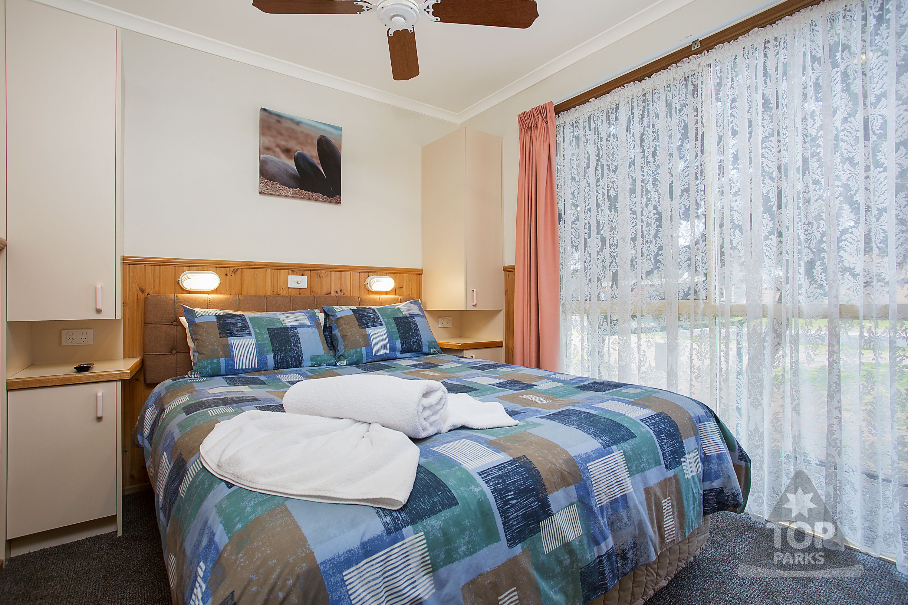 A hotel room with a large bed, a ceiling fan, a nightstand, and a window with sheer curtains.