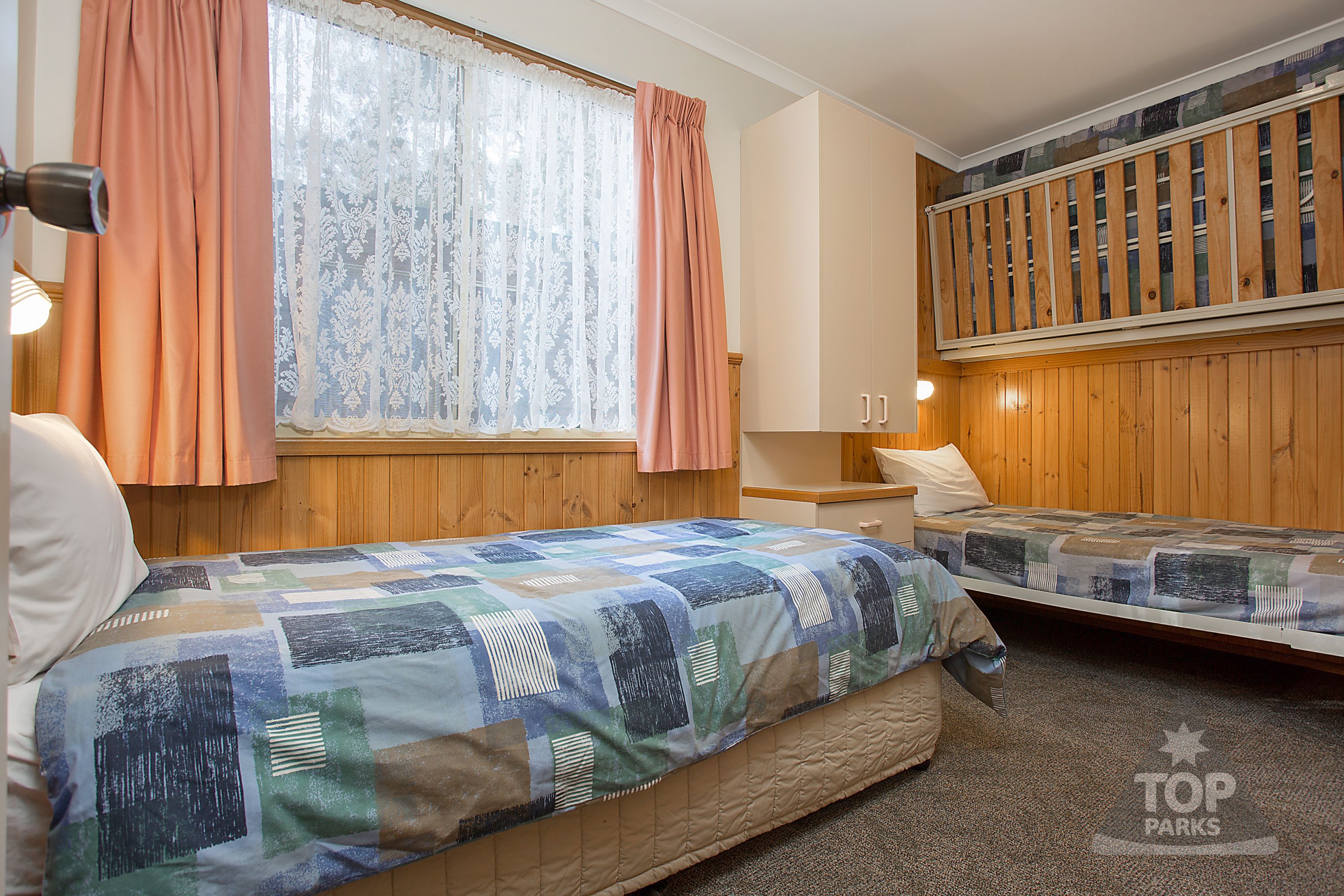 A bedroom with two beds, a window with curtains, a wooden wardrobe, and a nightstand.