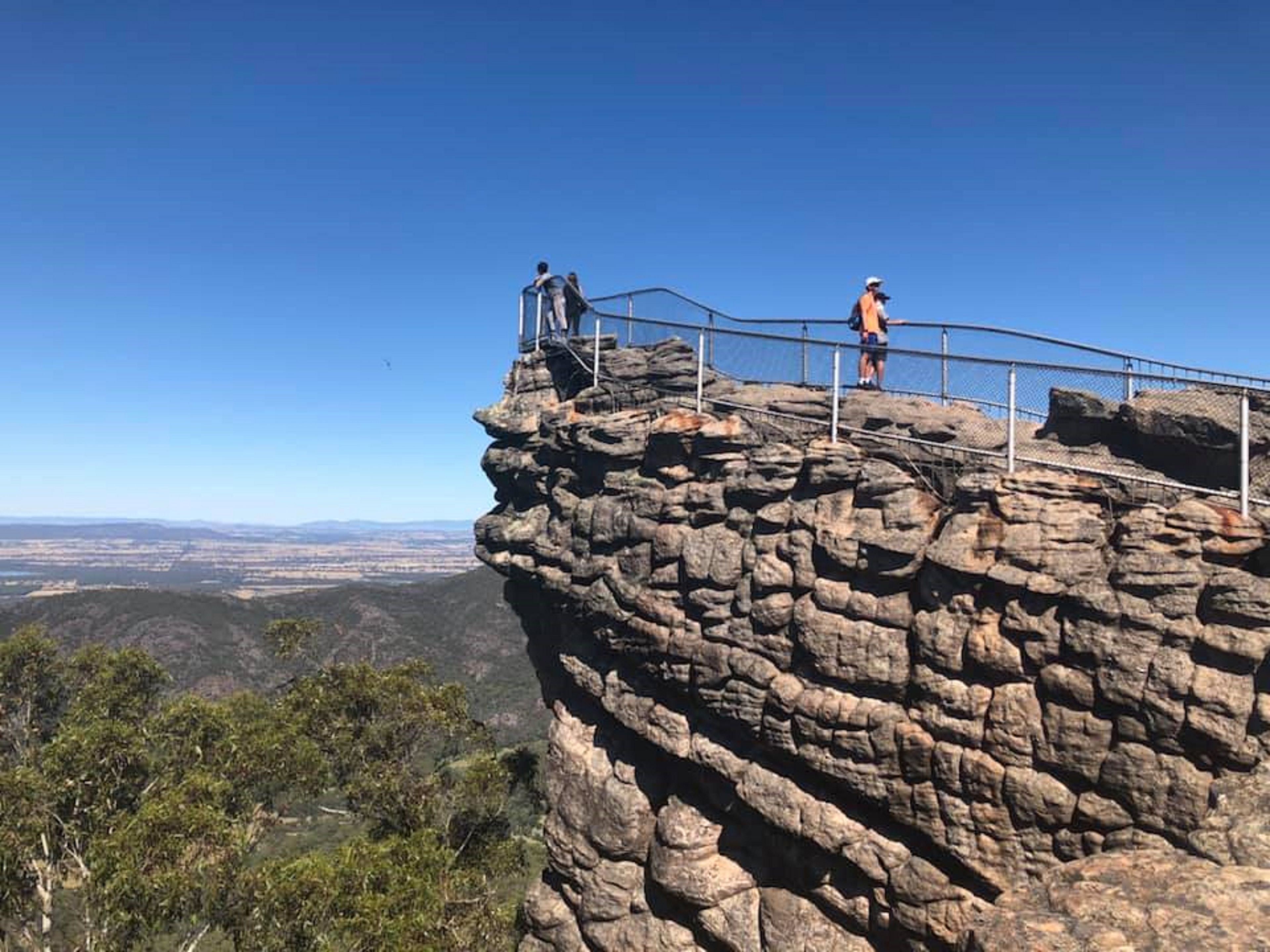 A rocky cliff with a metal railing and two people standing on top.
